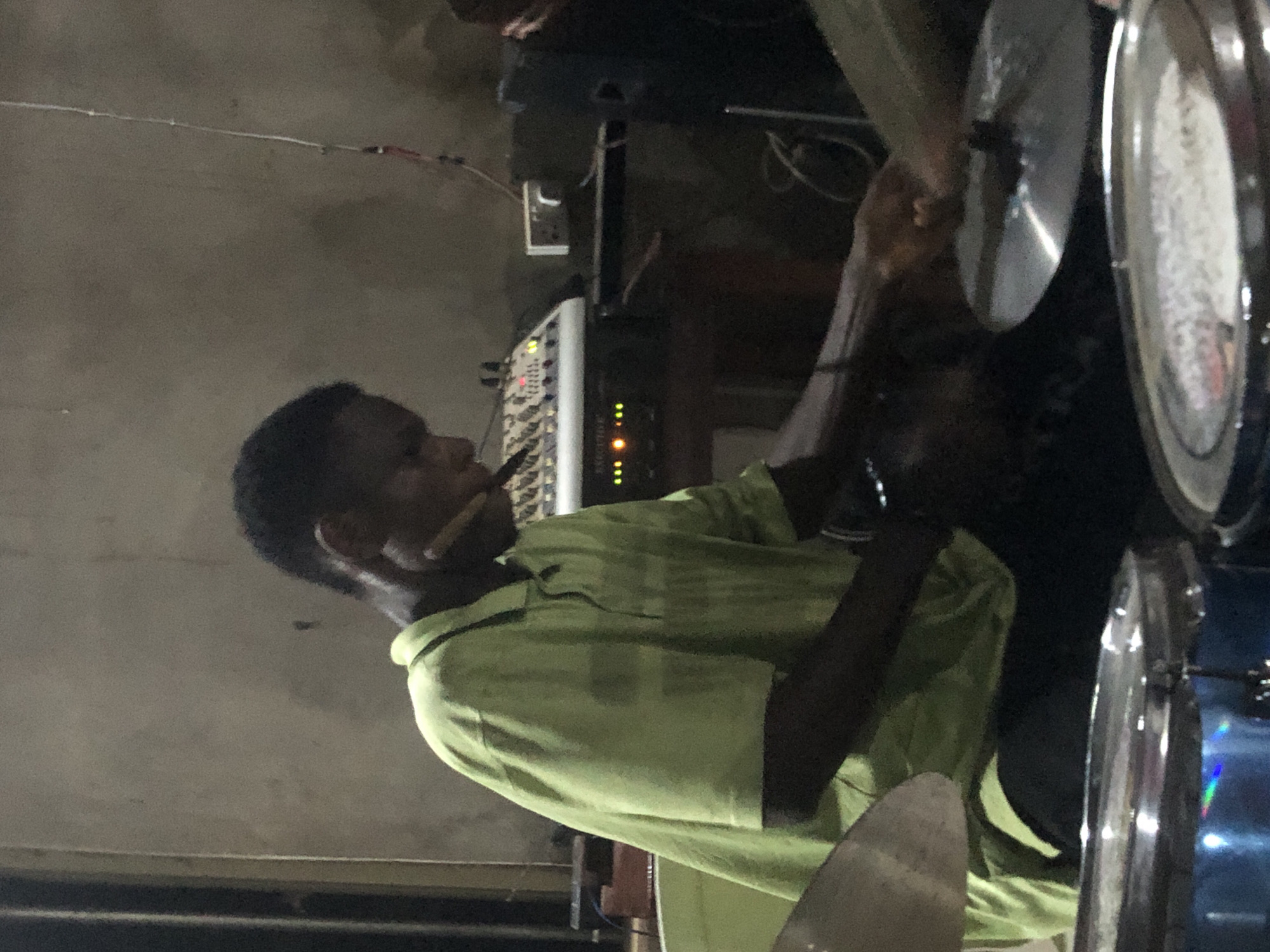 This vertically oriented image captures a dim indoor scene in Ukwu-Abwa, Nigeria, likely during the evening or night. The central subject is a young man, shown in profile, seated and actively playing a drum set. He wears a light green collared shirt and holds a drumstick, positioned as if striking one of the cymbals.
The drum set is partially visible, including a pair of cymbals to the left of the drummer's hands and another larger cymbal further back, which reflects some light. A blue drum is also discernible on the far right. To the left of the drummer, an audio mixer or soundboard is prominently displayed, featuring multiple white knobs, faders, and illuminated indicator lights in red, green, and yellow, signifying it is powered on. Below the mixer, a black electronic unit also shows active orange and green lights. An electrical outlet is visible on the wall behind the equipment, and various cables run across the scene, connecting the devices and stretching across the upper background.
The setting appears to be a functional space, possibly a rehearsal room, a small performance venue, or a church, equipped for musical performance or sound management. The background is dark and indistinct, suggesting a plain wall or ceiling. Another person's head is partially visible in the upper right corner. The ambiance is focused on the musical activity. No clearly legible text is discernible on the equipment, though a faint word resembling "MCATIVE" might be present on the black unit.