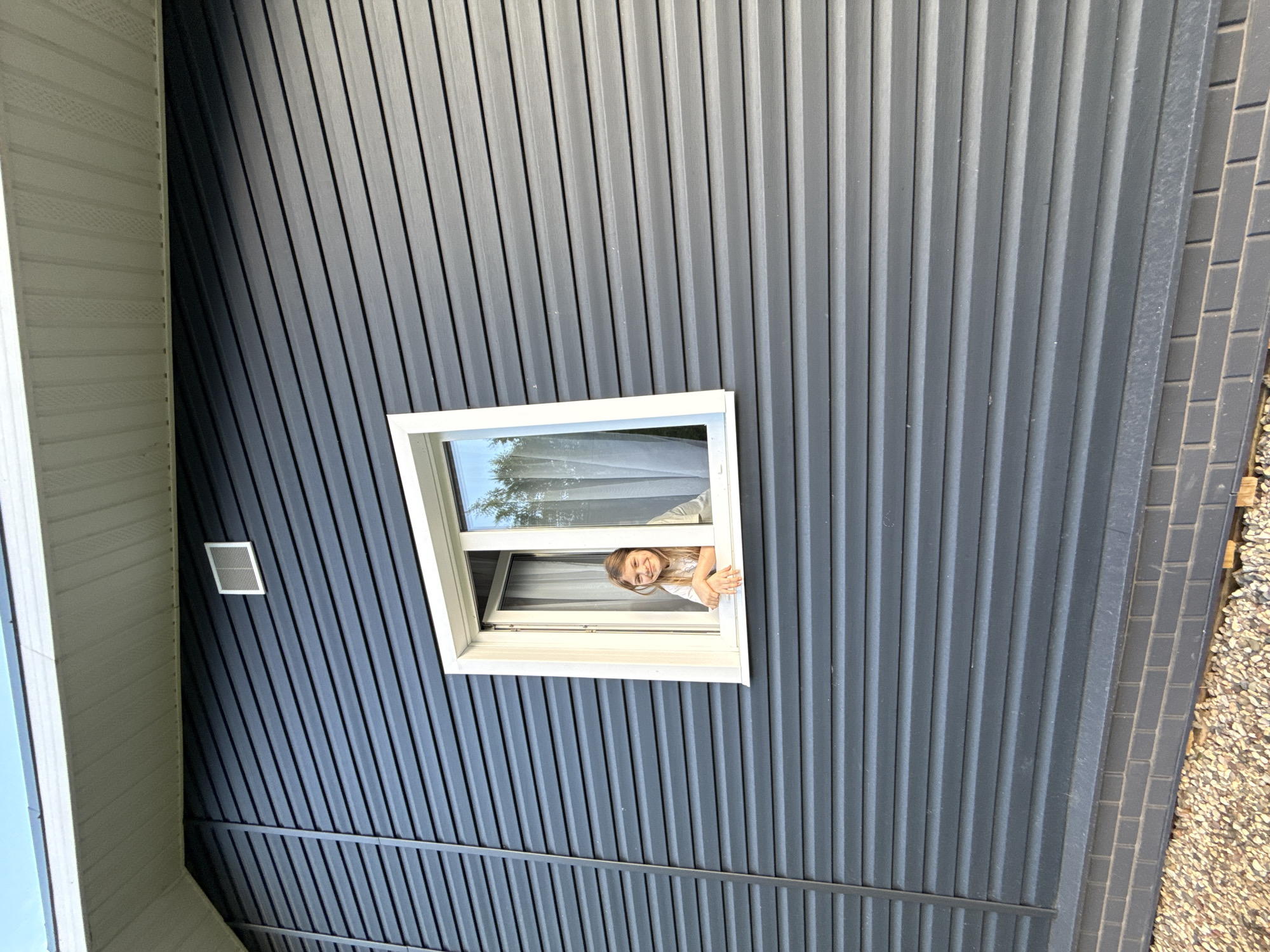 A young girl peers out of a white-framed window in a dark gray, corrugated metal building. The exterior of the building also has light gray elements and brick detail. The image was taken in Buzhaninovo, Russia.