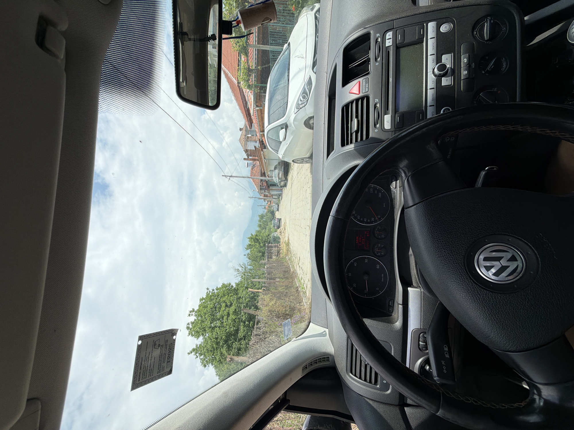 The image displays a first-person perspective from within a parked Volkswagen vehicle, looking forward through the windshield onto a street in Dabene, Bulgaria.
The interior of the vehicle shows a black leather-wrapped steering wheel with a chrome Volkswagen emblem at its center. Behind the steering wheel, the instrument cluster is visible, displaying "12:14" (time), "27.0°C" (temperature), and "570" on its digital readout. The center console features a silver-trimmed dashboard with a radio/navigation unit, climate controls labeled "CLIMATIC," and a red triangular hazard light button. A light-colored headliner is visible above, and a rearview mirror is centrally positioned, from which a small, cylindrical object resembling an air freshener hangs. A white sticker with black text, including "WICHTIG / IMPORTANT" and "2025," is affixed to the upper left portion of the windshield.
The exterior scene reveals an unpaved or cobblestone street stretching into the distance under a partly cloudy sky. On the right side of the road, a white Hyundai i30 hatchback is parked, facing the camera. Behind the Hyundai, multiple single-story houses with orange or red tile roofs are visible. On the left side of the street, a rustic fence composed of wooden posts is covered with dense green vegetation. Large green trees rise behind the fence, and further down the road, additional trees and utility poles with overhead wires are present. Distant mountains or hills are faintly visible on the horizon. Two other dark-colored vehicles are discernible in the distance along the street. No individuals are present in the observable frame.