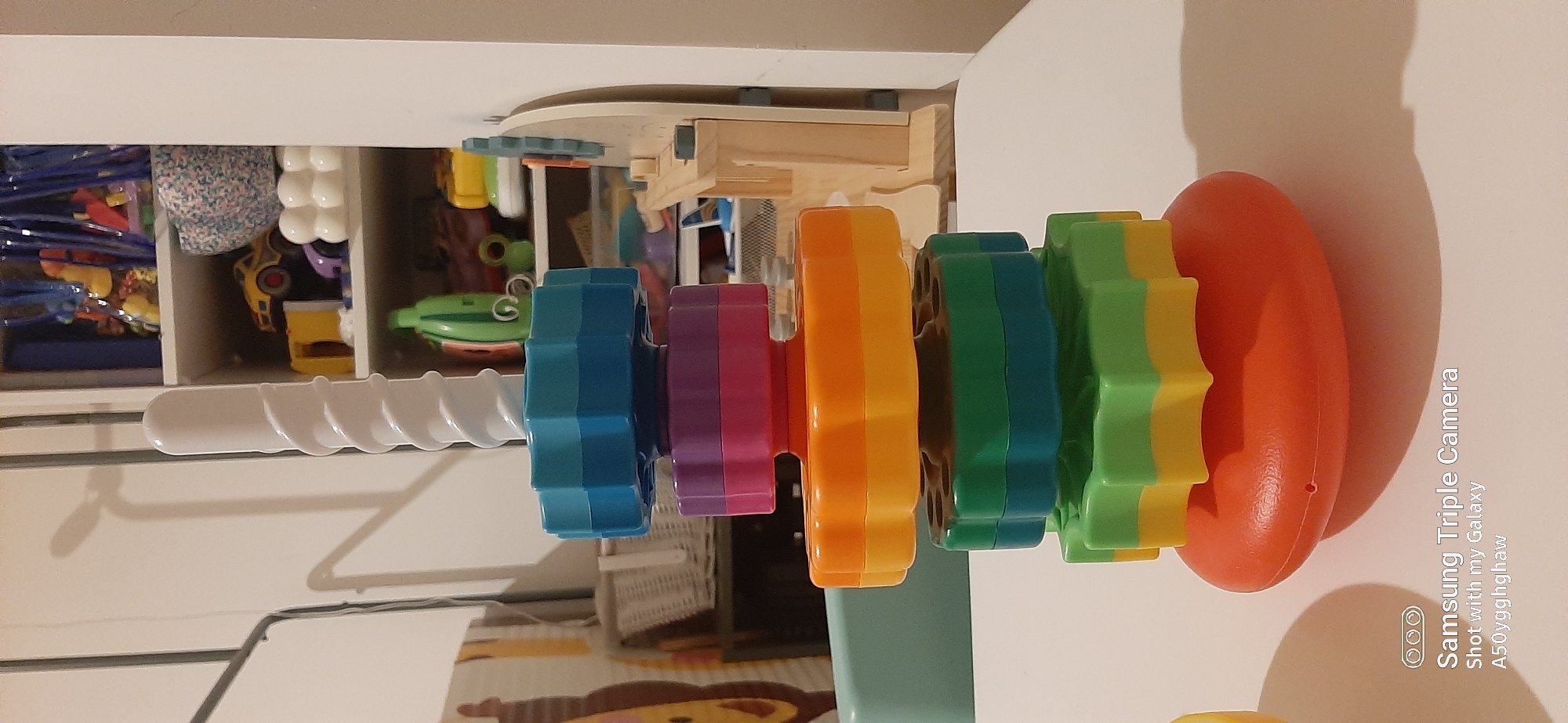 The image shows a colorful stacking toy with star-shaped rings on a white surface. The toy is predominantly orange, red, pink, purple, blue, and green. In the background, there are shelves filled with various toys and objects, including toy cars, containers, and a green bottle. A woven basket is visible to the left. The lighting suggests an indoor setting, possibly a home or playroom. The bottom of the image displays a watermark indicating "Samsung Triple Camera" and "Shot with my Galaxy A50yqgghghaw". There are no discernible elements indicating the location is in Dubai, United Arab Emirates, or any specific city.