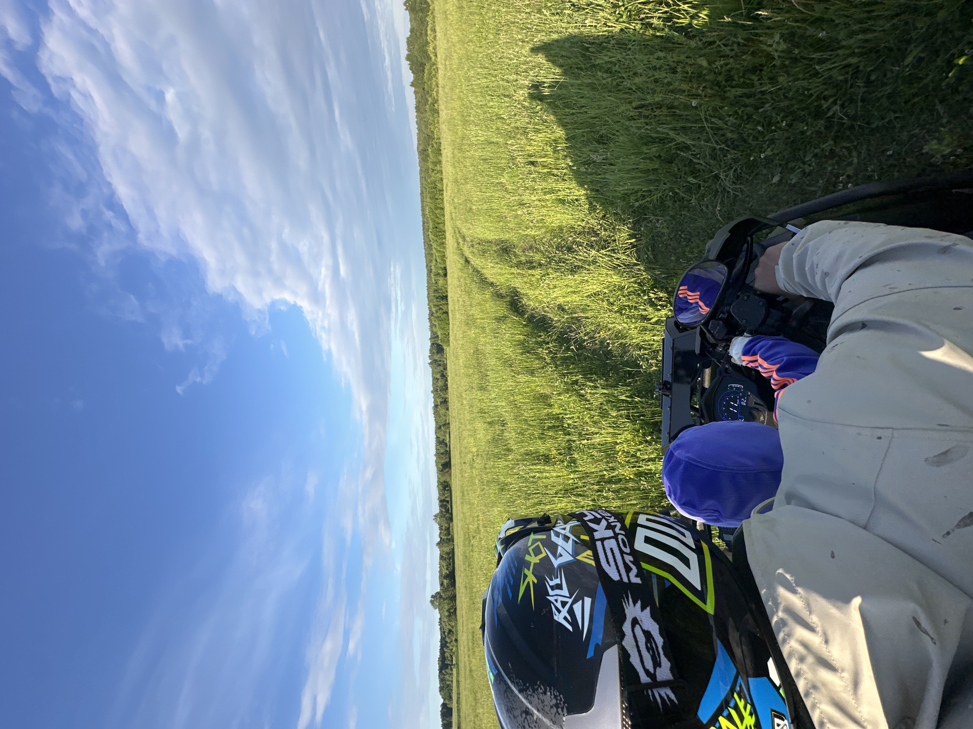 A first-person view from an All-Terrain Vehicle (ATV) moving through a grassy field in Kharnavy, Russia, under a partly cloudy blue sky. The upper third of the image shows a vibrant blue sky with scattered white clouds. The middle ground and foreground are dominated by tall, green grass, with a distinct two-track dirt path traversing through it, curving slightly to the right. A dense line of trees forms the horizon.
On the left side of the frame, the rear of a multi-colored motocross-style helmet is visible, featuring a black base with blue, yellow, white, and green graphic elements, including text such as "SHOT," "RACE GEAR," "SKY MONKEY," and the number "100." Below the helmet, the back of a person wearing a light-colored, possibly off-white or light grey, long-sleeved jacket or hoodie with visible dirt marks is present. A royal blue hood or cap is worn beneath the helmet. The person's right arm, clad in an orange-striped glove, extends forward to grip the right handlebar of the ATV. A digital dashboard to the right of the helmet displays "82." and other illuminated segments. A rearview mirror on the ATV reflects orange and blue fabric. Elongated shadows consistent with two human figures are cast forward and to the right onto the grass.