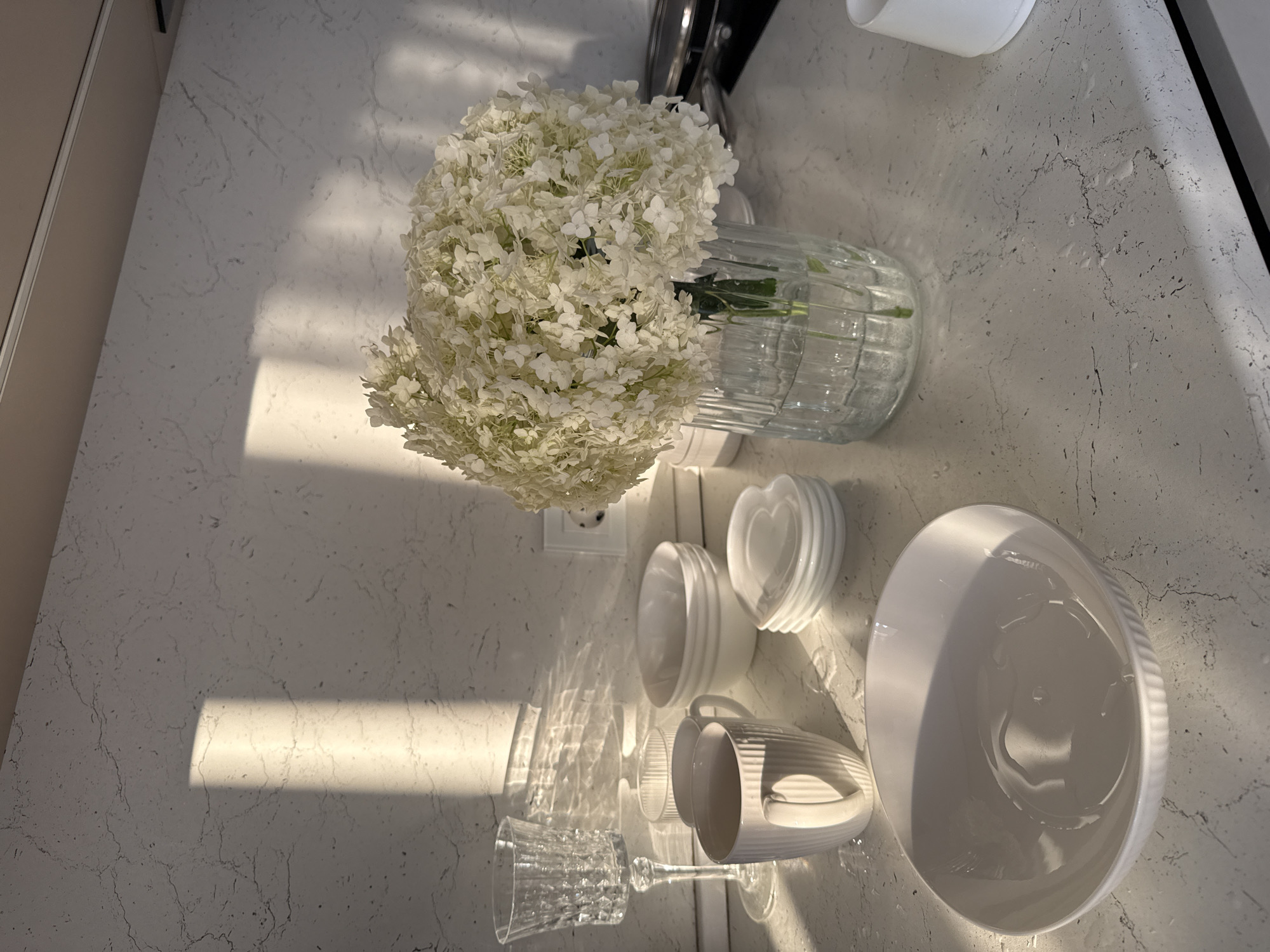 A close-up, top-down shot captures a white hydrangea bouquet in a ribbed glass vase placed on a white quartz countertop. The vase, with its ribbed texture, sits to the right of the flowers. The countertop has subtle grey marbling and is illuminated by a strong shaft of sunlight streaming from the left, casting a bright rectangular shadow across the surface.
Scattered around the vase and flowers are several white ceramic objects: a stack of nested bowls with ribbed exteriors, a heart-shaped dish, and a ridged mug. To the left of the mug, a clear, faceted glass goblet stands upright. The overall aesthetic is clean and bright, with a focus on white elements and the interplay of light and shadow. The setting appears to be a kitchen or dining area, possibly a countertop, given the presence of decorative and functional ceramicware. The sunlight suggests it is daytime, likely morning or afternoon, depending on the orientation of the light source. No people are visible, and there is no discernible text in the image. The scene evokes a sense of calm and order, enhanced by the natural light.