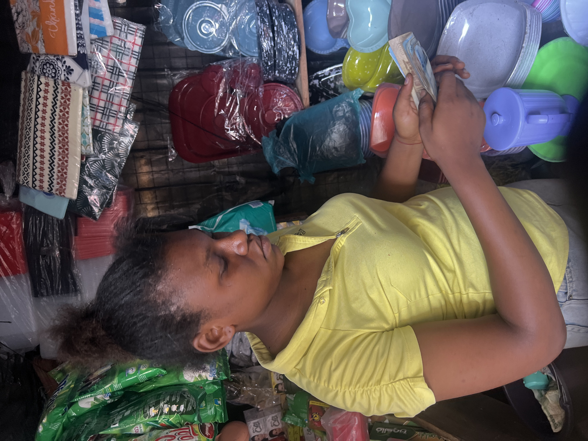The image shows a woman in Nkpor, Nigeria, sitting amidst a variety of items for sale in what appears to be a small retail space or stall. The woman, wearing a yellow top, is counting Nigerian Naira banknotes in her hands. Around her are various household goods, including stacks of disposable plates and containers in different colors, dish towels, and packages of what seems to be food or detergent. The items are arranged in close proximity, suggesting a limited space for displaying the goods. The overall environment appears to be an informal market setting.
