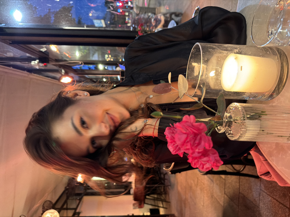 The image depicts a young adult female seated at a table inside a restaurant or cafe in Tel-Aviv, Israel, during evening hours. The woman, with brown hair, looks directly at the camera with a subtle smile, her left hand gently touching her cheek. She wears a black, collared, long-sleeved top with a visible zipper, stud earrings, a delicate necklace with two pendants, and a red string bracelet on her left wrist, alongside another bracelet featuring a small, circular, reflective element. A faint tattoo is visible on her inner left forearm.
On the white-clad table, a clear ribbed glass vase holds three vibrant pink carnations and a single green stem. Adjacent to this, a large clear glass hurricane lamp encloses a lit white pillar candle, providing ambient light. A clear glass tumbler is partially visible to the right of the candle. A light-colored garment or bag is partially visible near the lower left edge of the table.
The background reveals the interior of the establishment with blurred dark furniture and warm lighting from spherical and reddish-orange wall-mounted fixtures. Beyond a large glass window, the urban environment under a dark blue sky is visible, characterized by distant city lights, blurred red and green traffic lights, and reflections of street activity.