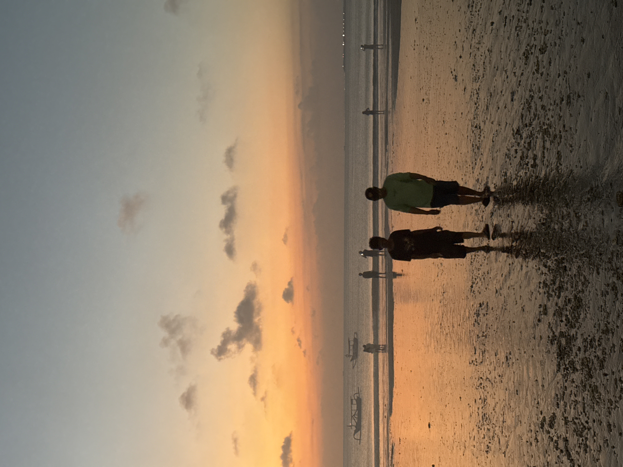 At sunset in Kuta Selatan, Indonesia, two men walk on a wet beach. The sky transitions from a pale blue to an orange hue, with scattered clouds. The ocean reflects the sky's colors. Several boats are visible on the water. Other people are scattered along the shoreline, some standing in the water. The sand is speckled with small shells and debris.