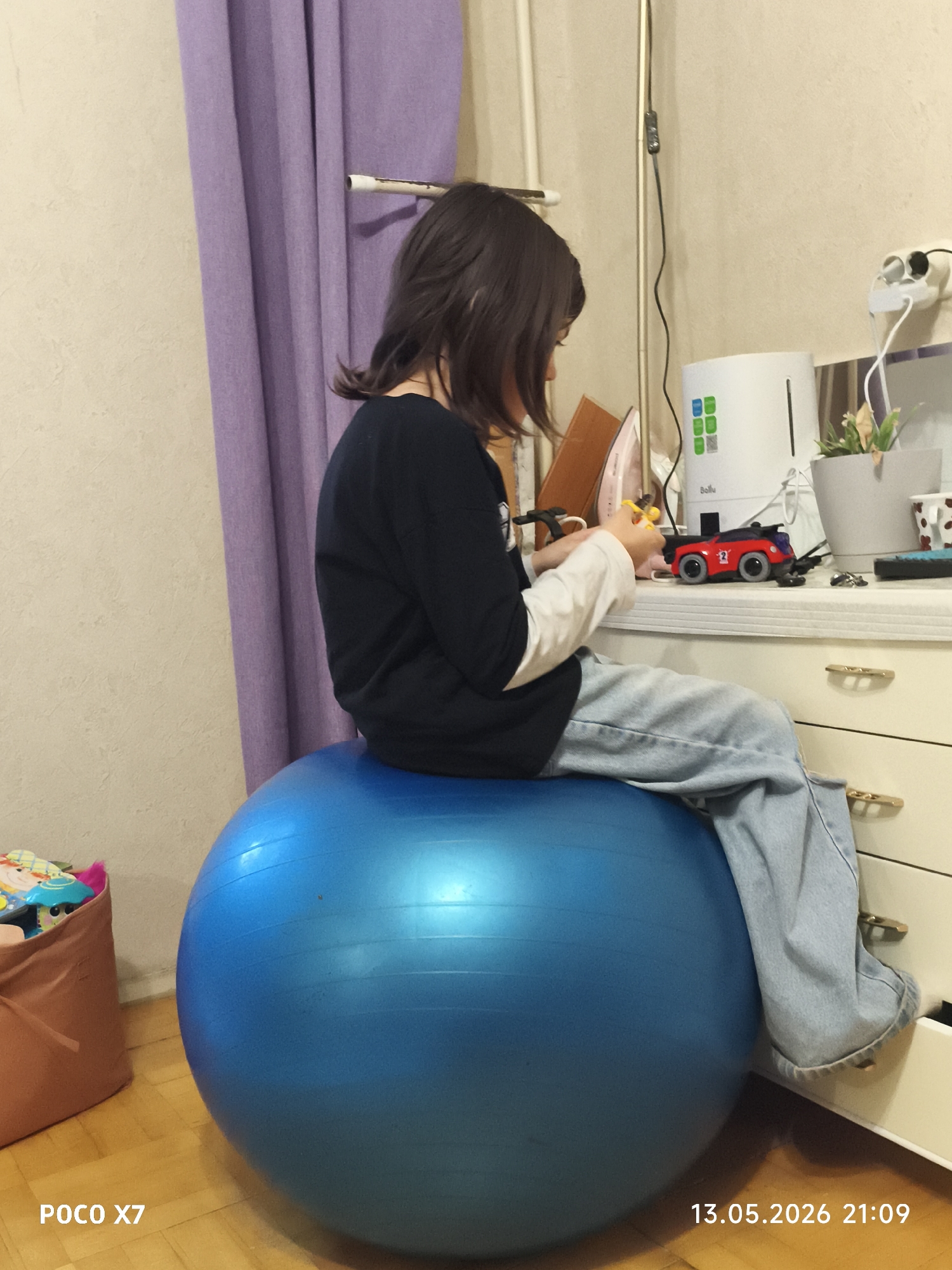 A child sits on a large blue exercise ball in a room with beige walls, light purple curtains, and wooden flooring. The child wears a dark long-sleeved shirt, a white undershirt, and light-wash jeans. The child is seated facing a white dresser with brass-colored hardware, and appears to be holding and manipulating small yellow and black plastic pieces. On the dresser sits a red and black toy car with the number "2" on its side, a humidifier, an open book, and a potted plant. A brown tote bag with toys is partially visible next to the ball. The camera watermark indicates the image was taken with a POCO X7, and includes the date 13.05.2026 21:09. While the location is specified as Chaikovsky, Russia, the image only shows the interior of a room.