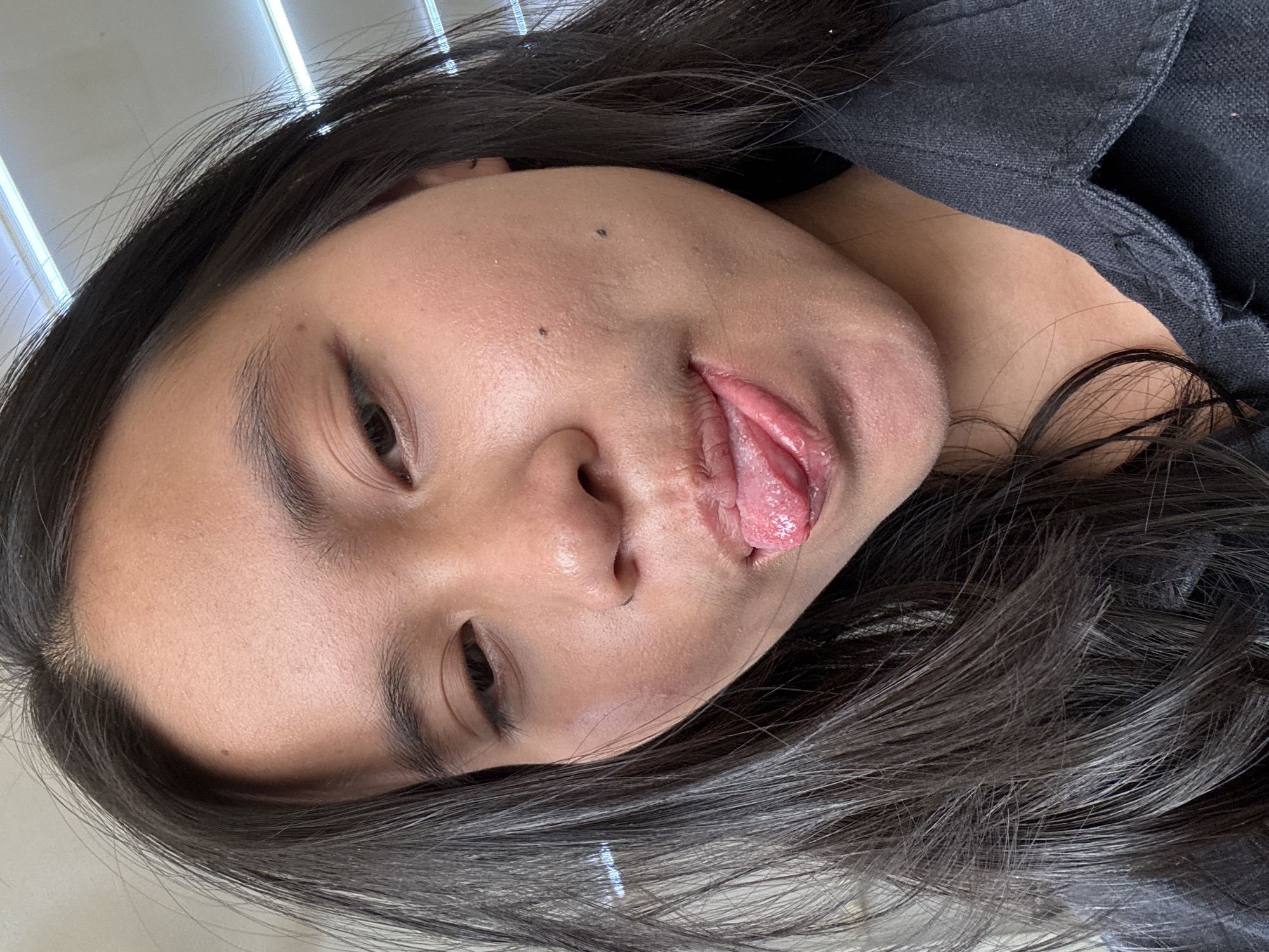 A close-up, selfie-style, medium-shot photograph depicts a young East Asian individual with long, dark hair. The person's face is angled slightly down and to their left. Their tongue is extended, with the tip touching their lower lip, and the mouth is partially open. The skin on their face appears to have a slight sheen. Two dark moles are visible on the right side of their face. They are wearing a dark-colored shirt. The background is out of focus and appears to be an indoor setting with fluorescent lighting visible at the top of the frame. The image was taken in East Jakarta, Indonesia.