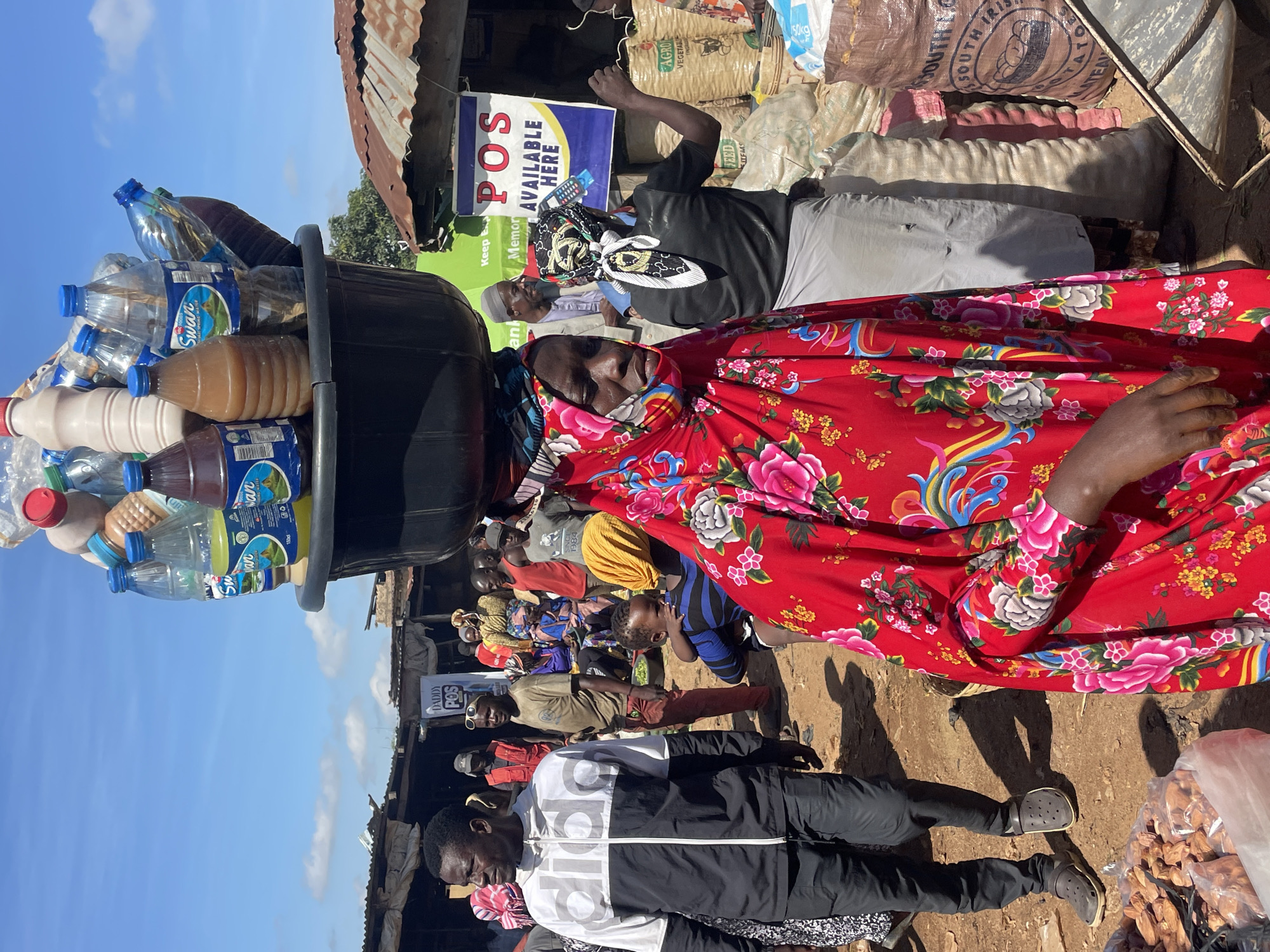 In Gura Top, Nigeria, a crowded market scene unfolds under a bright, clear sky with scattered clouds. The central figure is a woman wearing a vibrant red floral-patterned dress and a headscarf, balancing a large black basin filled with various bottles of drinks on her head. The bottles appear to be water, juice, and possibly milk-based beverages, some with blue and white labels.
To the left, a man dressed in a black and white Adidas jacket and black pants walks away from the camera, his head turned to the side. He is wearing grey Crocs. In the background, other market-goers mill about, some standing, others sitting. A man in a brown t-shirt and red pants stands near a market stall, and another man wearing a light grey suit and a white skullcap is visible behind the woman with the basin.
Market stalls with corrugated metal roofs are scattered throughout the scene. One stall has a sign that reads "POS AVAILABLE HERE." Bags of produce, possibly potatoes judging by the text on one bag, are stacked behind some of the stalls. The ground is dirt, indicative of an outdoor market. The overall atmosphere appears busy and dynamic.