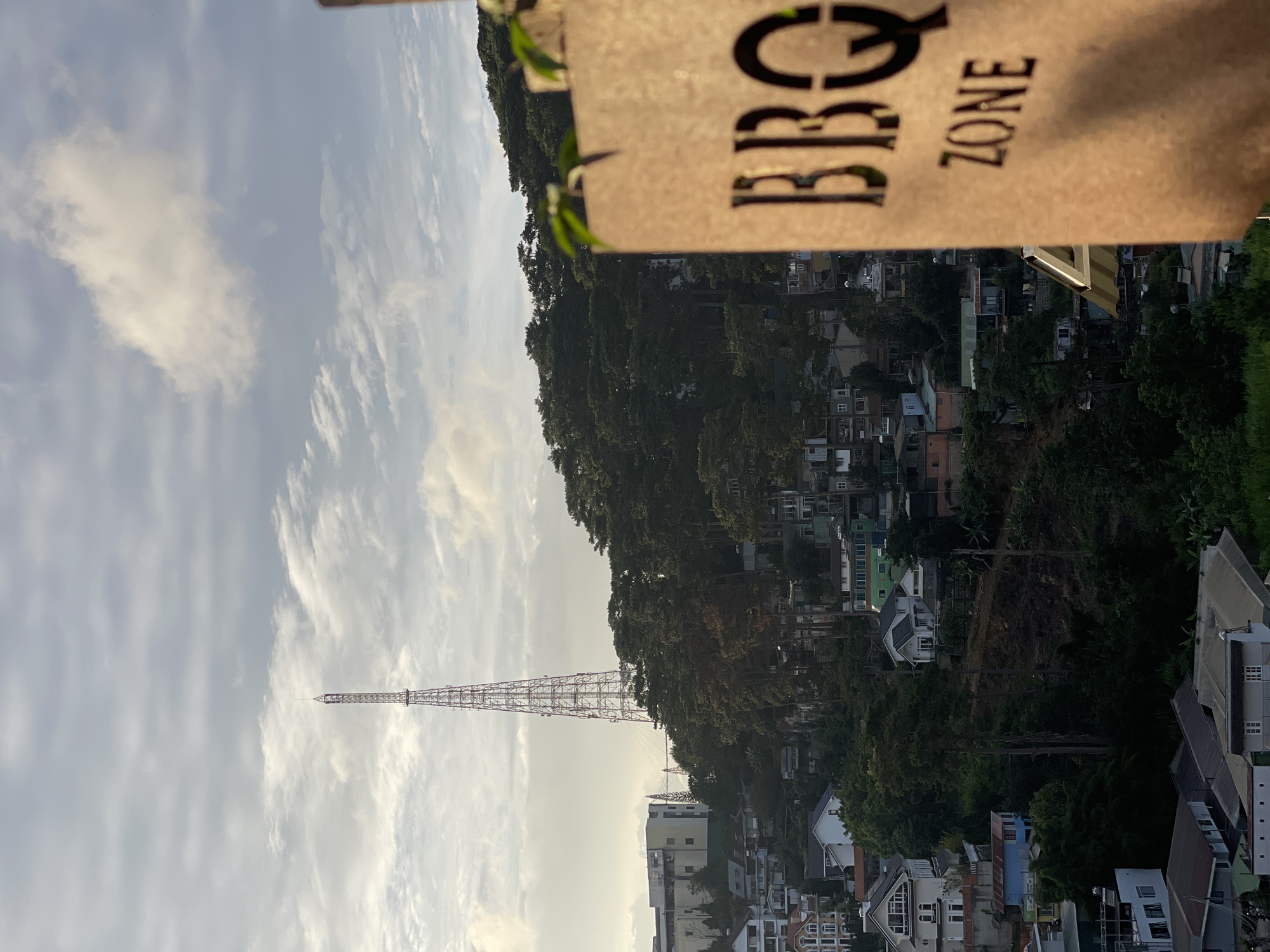 The image depicts an elevated view of a hillside city, likely Da Lat, Vietnam, characterized by a dense scattering of buildings and abundant greenery. A prominent telecommunications tower stands tall on a distant hill, piercing the partly cloudy sky. In the foreground, a rustic, wooden sign with cut-out letters reads "BBQ ZONE," partially obscuring the cityscape. The architecture of the buildings appears varied, with some modern structures and older, more traditional homes. Lush trees and foliage are integrated throughout the urban landscape, suggesting the city's natural setting. The lighting indicates either early morning or late afternoon, with soft, warm sunlight illuminating parts of the scene. No individuals or explicit actions are visible.