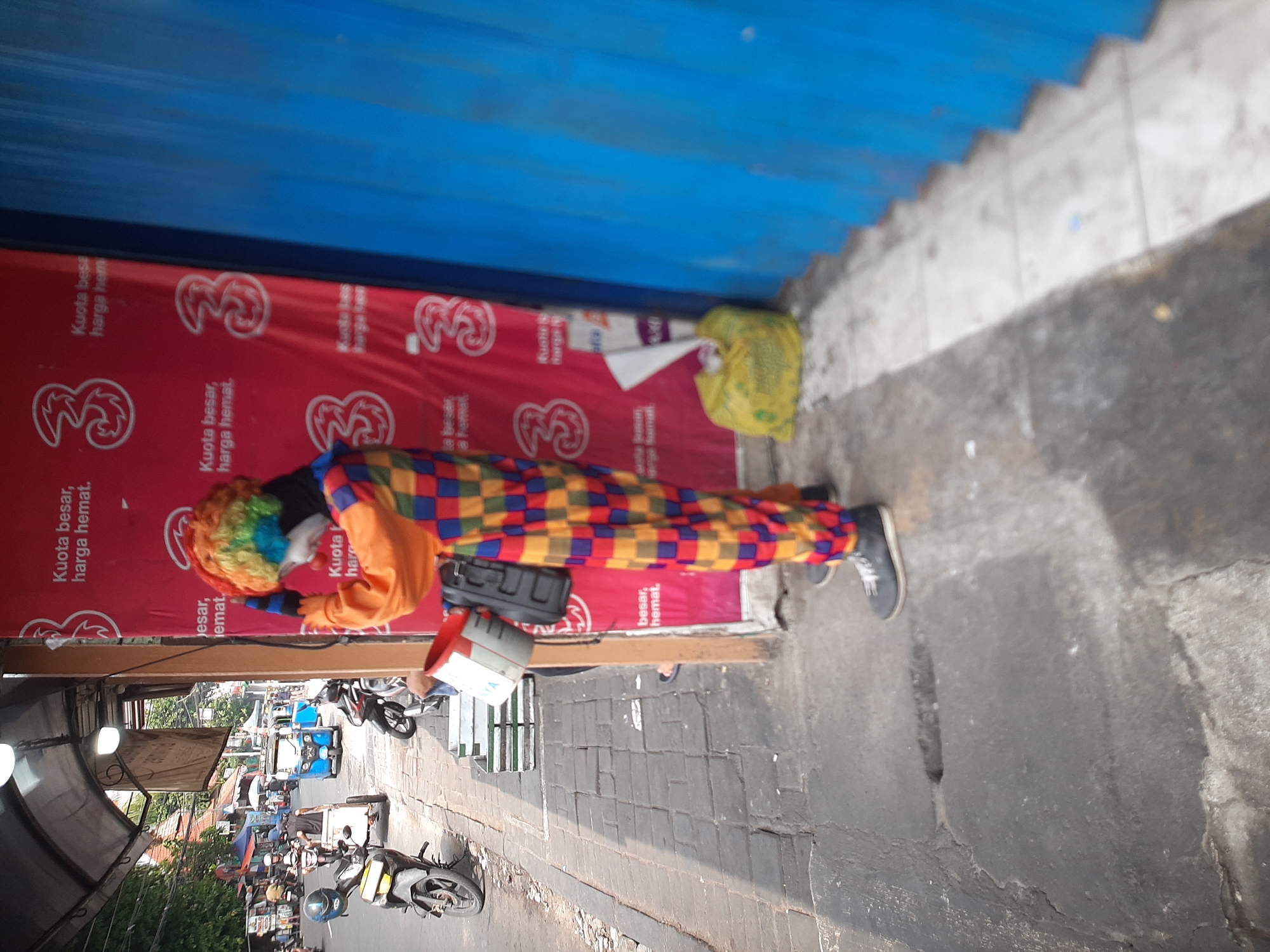 In Central Jakarta, Indonesia, a person dressed as a clown stands on a sidewalk next to a building with a red banner displaying "Kuota besar, harga hemat." The clown wears a colorful wig with red, yellow, blue, and green hues, and a multi-colored checkered costume with orange, red, yellow, and blue squares. Their face appears to be painted white with red and blue accents. The clown is holding a silver bucket with a black handle and a black strap is visible over their shoulder. To the right of the clown, a yellow plastic bag lies on the ground, and a blue corrugated metal structure is present. In the background, several motorcycles are parked, and a blue three-wheeled vehicle is visible on the street. The sidewalk is made of grey bricks, and there is a prominent crack in the pavement in the foreground. The scene appears to be a street in a bustling urban area.