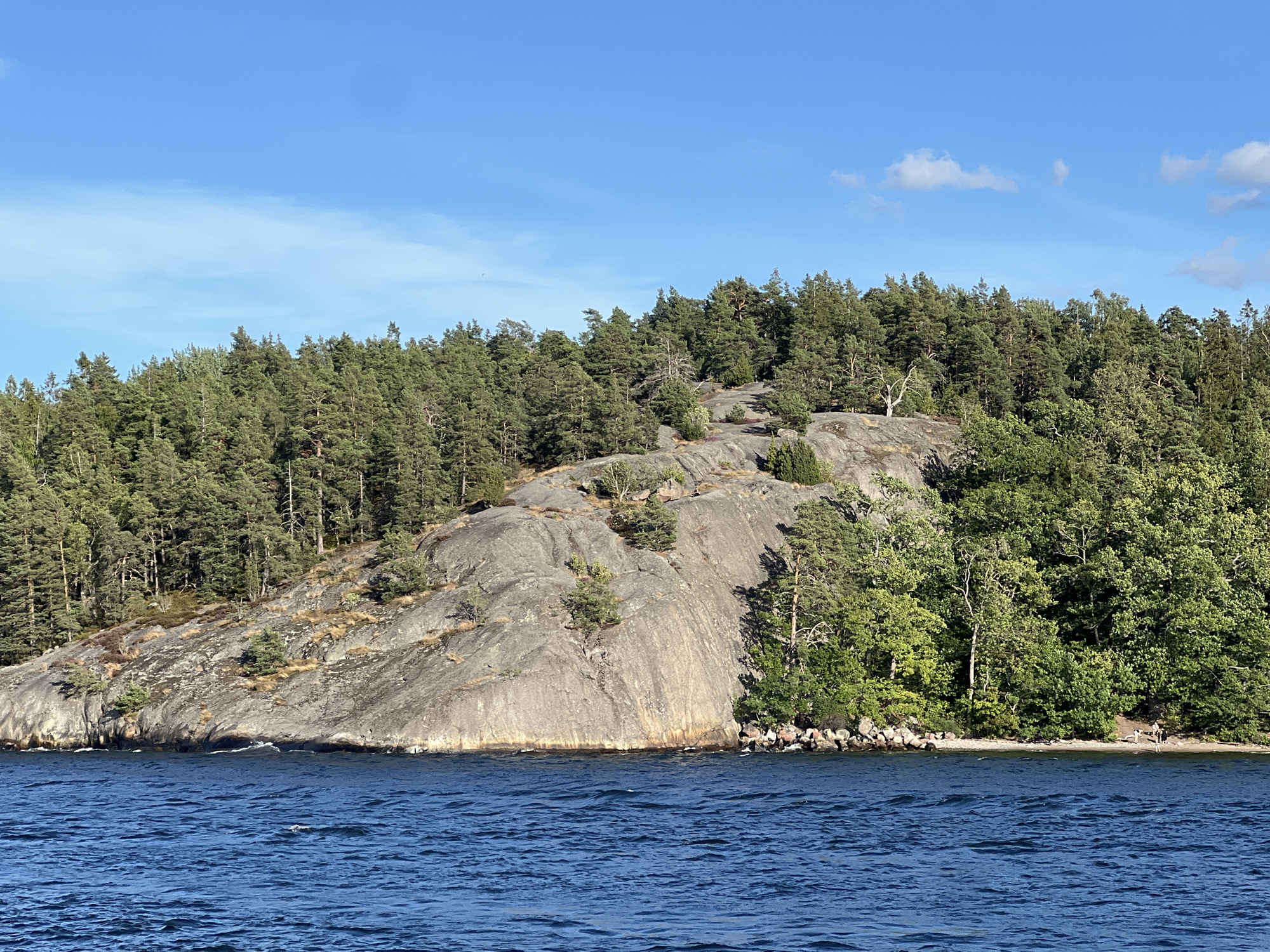 The image depicts a scenic view of a rocky island covered with lush green coniferous trees. The island is situated in the blue waters of the Baltic Sea, likely within the archipelago near Lidingö, Sweden. A rocky, uneven shoreline leads up to the densely wooded slopes. The sky above is a clear blue with a few wispy white clouds. In the foreground, the water is a deep blue with gentle ripples. On the far right of the shoreline, a small group of people can be seen on a rocky beach, suggesting a recreational area or a popular spot for swimming or picnicking. The overall impression is one of a serene, natural landscape.