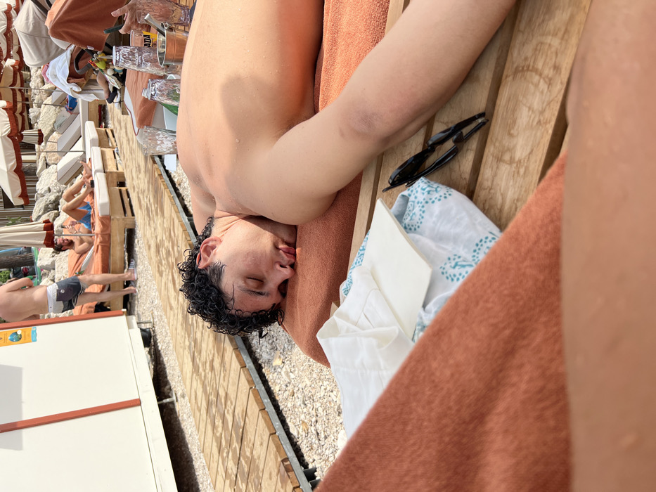 A man with dark curly hair sleeps on a towel-covered lounge chair. Sunglasses and a patterned white cloth rest beside him. A beach setting includes additional lounge chairs under orange umbrellas, some occupied by other people. A white building with an orange stripe is visible in the background, with light-colored gravel surrounding the chairs. The image is taken at an angle.