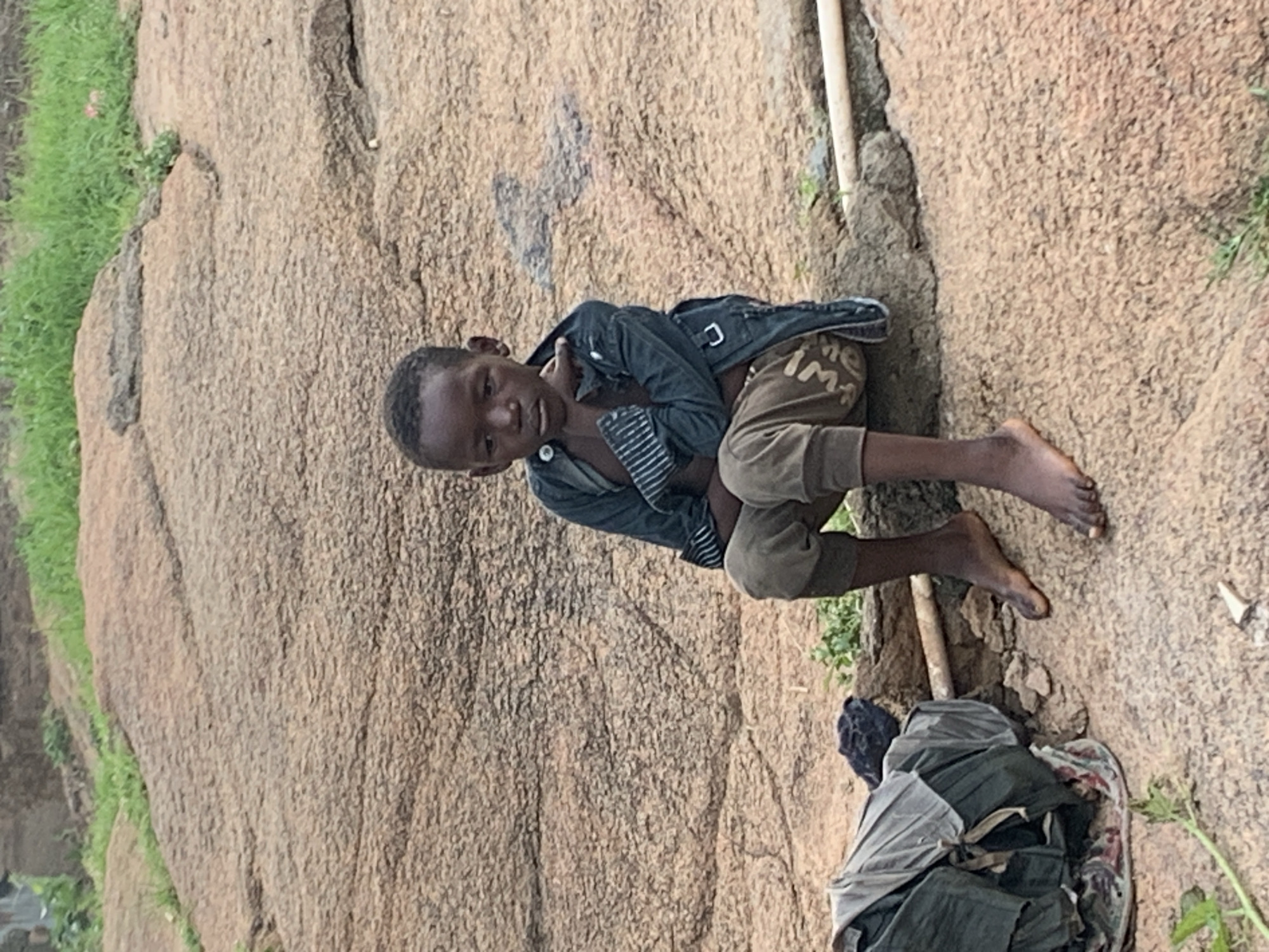 A young boy sits on a large, weathered rock formation, with a pile of dark clothing to his left. He is wearing a dark jacket over a lighter-colored shirt and khaki pants. His arms are crossed, and he gazes downward with a somber expression. The background shows more of the rocky terrain, with patches of dry grass and a hint of green vegetation further up the slope. The overall scene suggests a rural or semi-rural environment, possibly on the outskirts of Jos, Nigeria, given the characteristic geological features of the region.