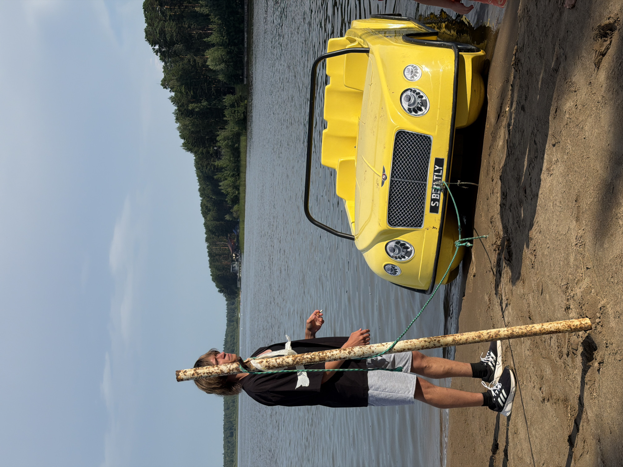 A man stands on a sandy shore next to a bright yellow pedal boat shaped like a Bentley. The boat is partially in the water. The man is wearing grey shorts, a black short-sleeved shirt over a white tank top, and black and white athletic shoes. He is holding a cigarette and a rusty pole is in front of him, tied to the boat with a green rope. The background shows a body of water, a tree-lined shore, and a cloudy sky. The location is the Kama River in Chaykovsky, Russia.