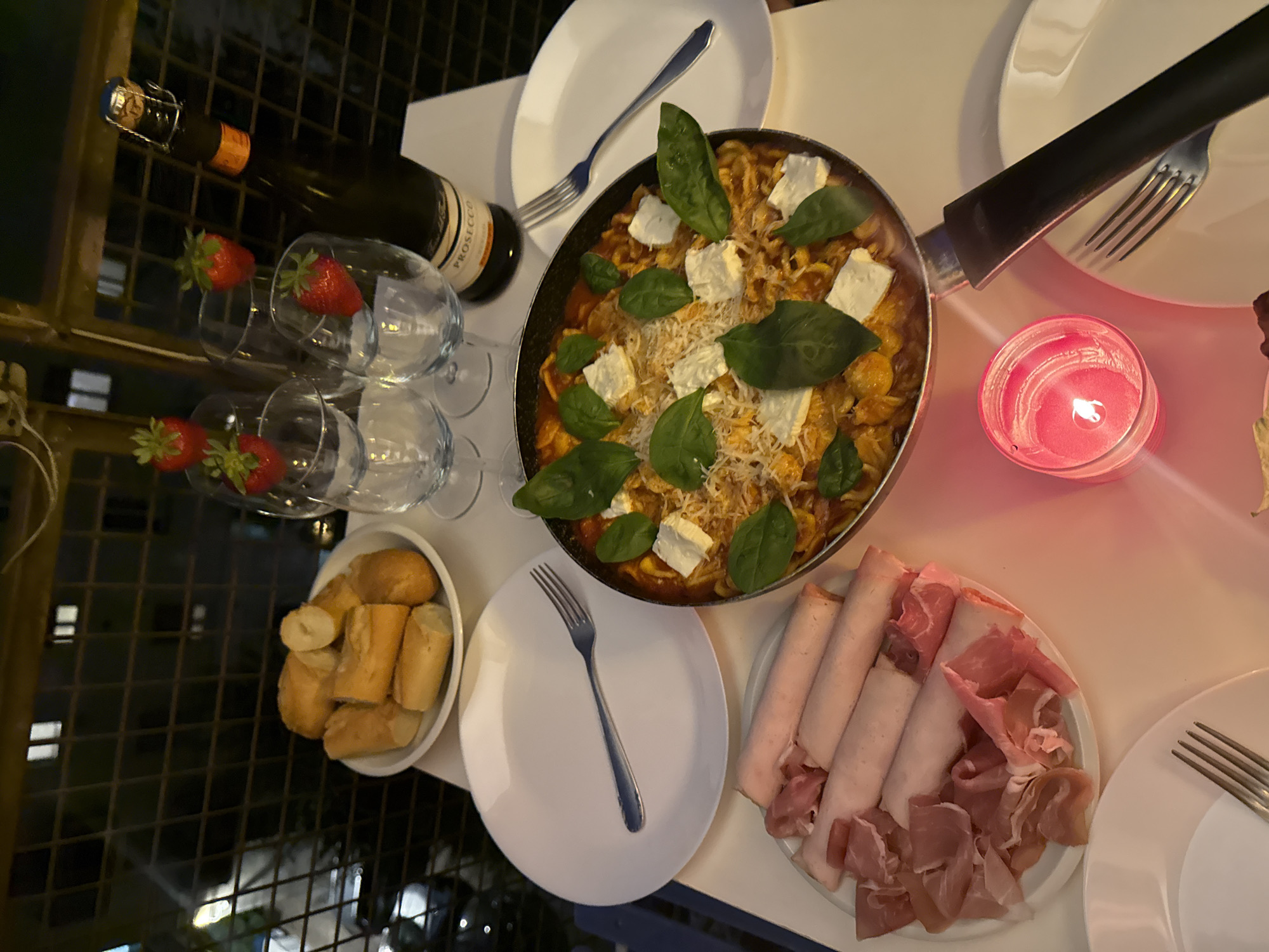The image presents an inviting and intimate outdoor dinner setup at night, likely on a balcony or patio in Milan, Italy. The scene is softly lit by a glowing red candle set in a glass holder, creating a warm and romantic ambiance.
The main culinary highlight is a large pan filled with pasta, possibly gnocchi or small shell pasta, coated in a rich red sauce. This dish is generously garnished with cubes of white cheese, grated cheese, and vibrant green basil leaves. Alongside, a white bowl holds several pieces of sliced baguette or similar bread. Another plate offers an assortment of cold cuts, featuring neatly rolled slices of light pink ham and delicate, crumpled slices of prosciutto.
For drinks, a bottle of sparkling wine, clearly labeled "PROSECCO," stands ready. Several stemmed wine glasses are elegantly arranged, each containing a fresh red strawberry, adding a touch of sweetness and visual appeal. White round plates and silver forks are laid out, suggesting a meal for at least two people.
The background shows a dark, grid-like structure, likely a balcony railing, and blurred lights of a city below, confirming an urban outdoor setting after dusk. The combination of food, drink, and candlelight evokes a cozy, relaxed, and special evening meal.