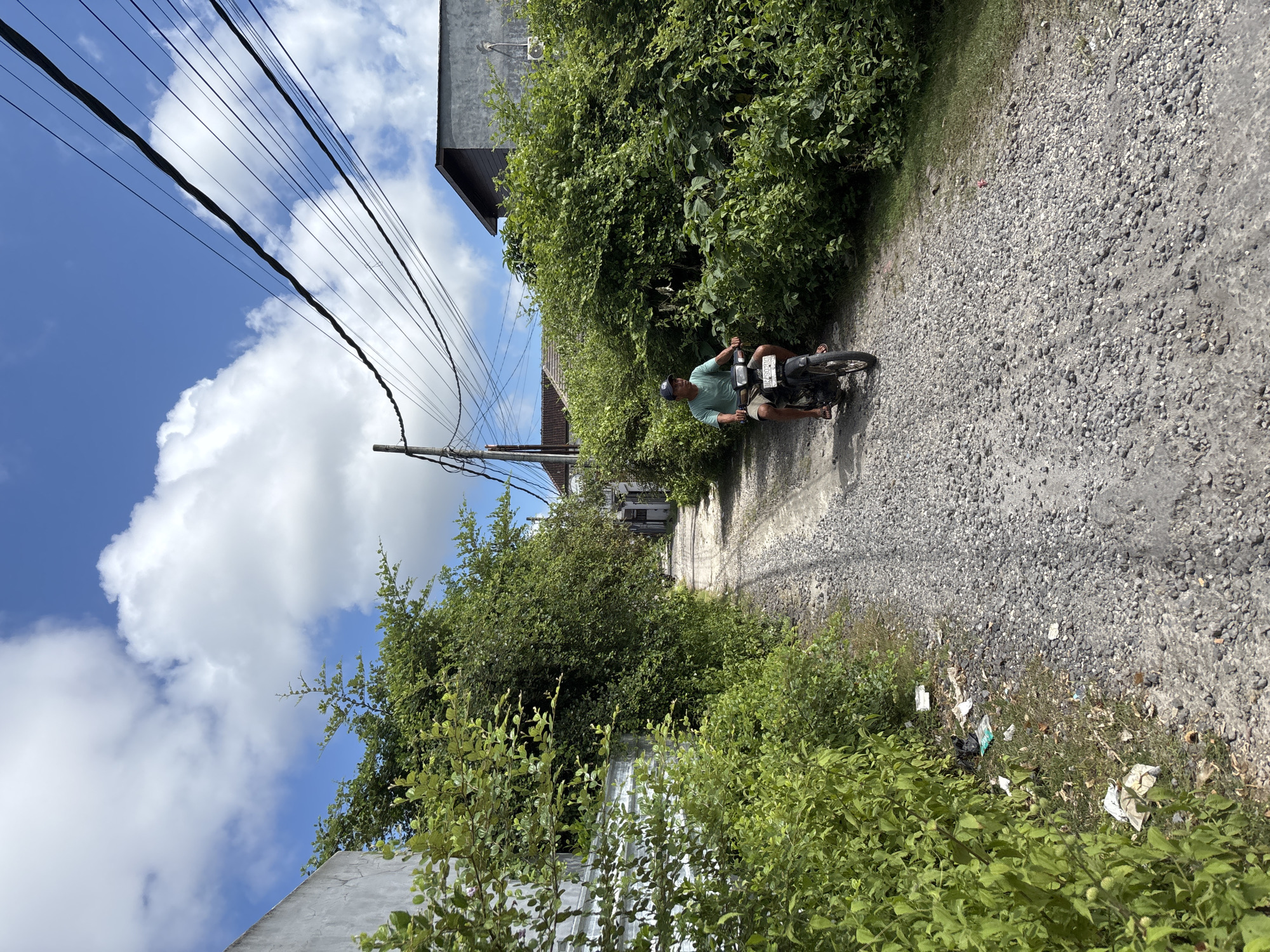 The image depicts a man riding a motorcycle on a gravel road in Kuta Selatan, Indonesia. The road is bordered by dense vegetation on both sides, with glimpses of buildings visible through the foliage. Power lines stretch across the top left of the frame, silhouetted against a blue sky with scattered clouds. Litter is visible along the roadside. The man is wearing a light-colored shirt, shorts, and a baseball cap, and he appears to be looking straight ahead.