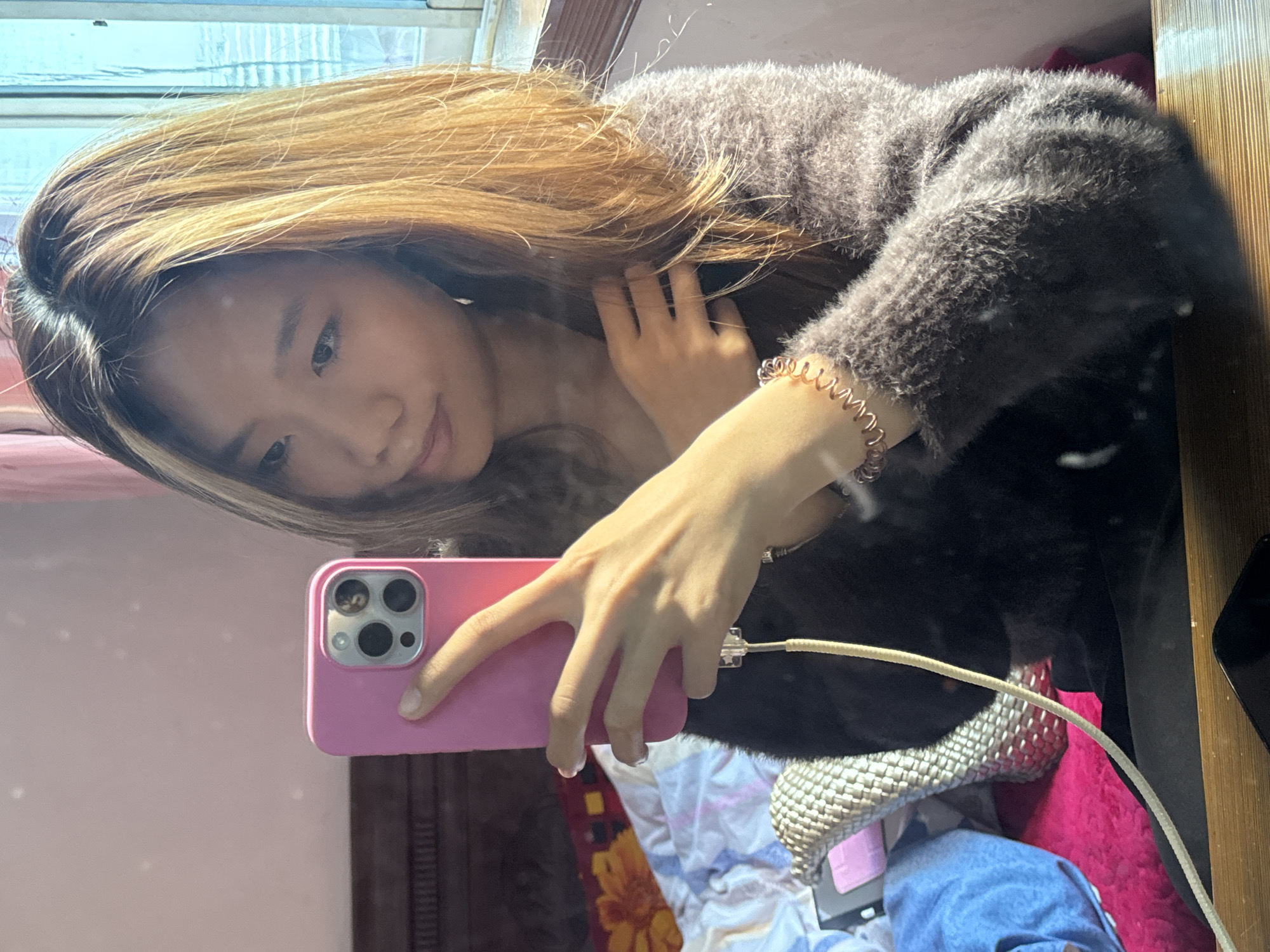 A young woman with blonde hair is taking a selfie in a mirror. She is wearing a dark, fuzzy sweater and holding a pink smartphone with a white charging cable plugged into it. The background shows a room with a window and a bed with patterned bedding. The location is possibly Hechuan, China.