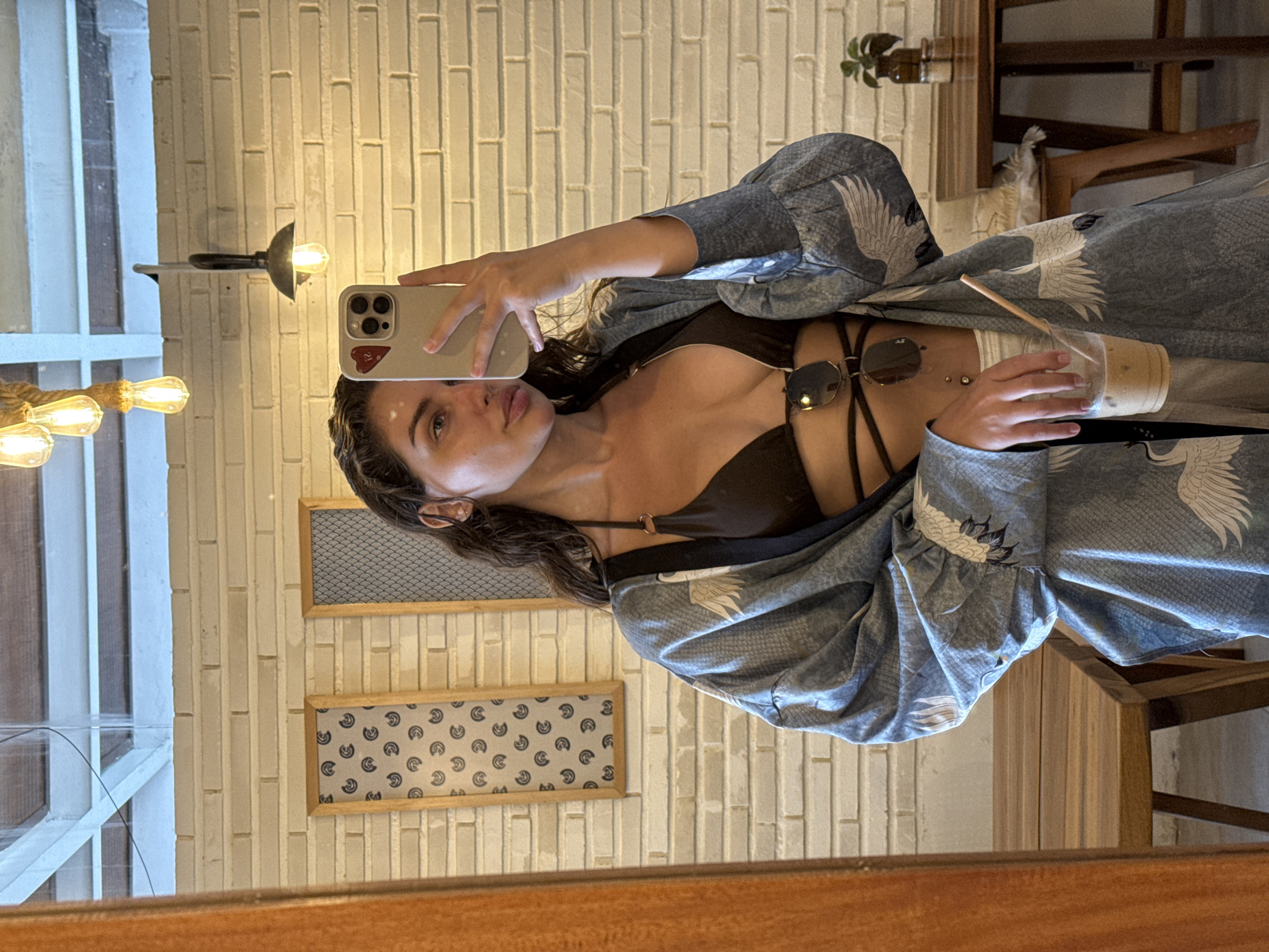 A light-skinned person wearing a black top, a blue and gray robe-like garment with a crane pattern, and sunglasses is taking a selfie with their iPhone in a cafe setting in the city of Unknown, Indonesia. They are holding a clear cup containing a beverage with a straw. Behind them, the wall is made of white bricks, and there are two framed patterned artworks. Part of the window and lighting fixture are visible on the left. On the right, there is a table with a plant and a partially visible chair. The bottom of the image shows a wooden frame.