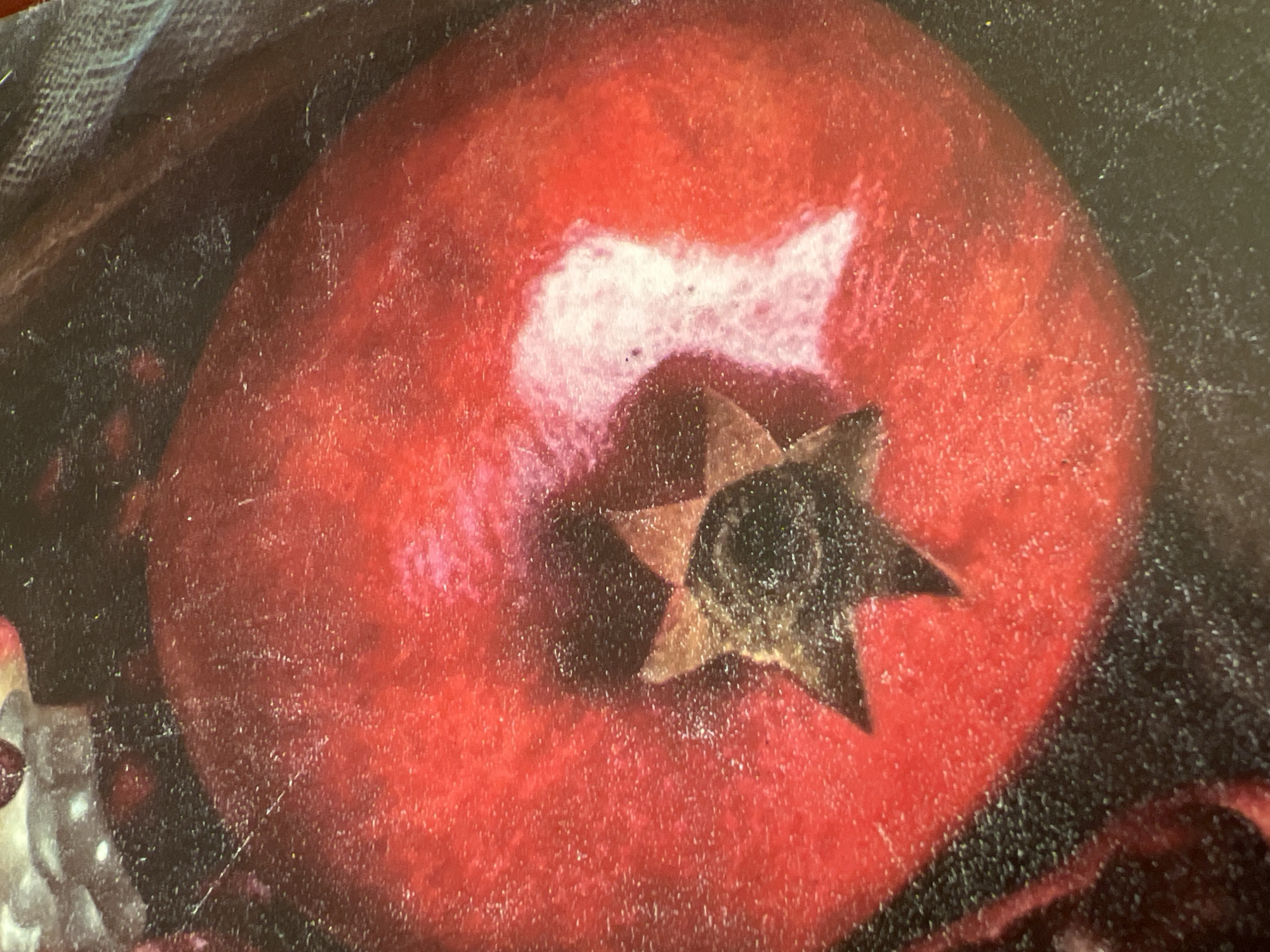 The image is a close-up, top-down view of a pomegranate. The fruit is a deep red, with a star-shaped calyx at the top. There is a highlight on the upper right side of the pomegranate, suggesting it is wet or has a glossy surface. The background is dark and out of focus, with some texture visible, possibly fabric or another surface. The image has a distressed, textured appearance, with visible scratches and dust particles. There is no indication of the city of Minsk, Belarus, or any environmental context beyond the immediate subject. The image depicts a single object, a pomegranate, and no people or actions are present.