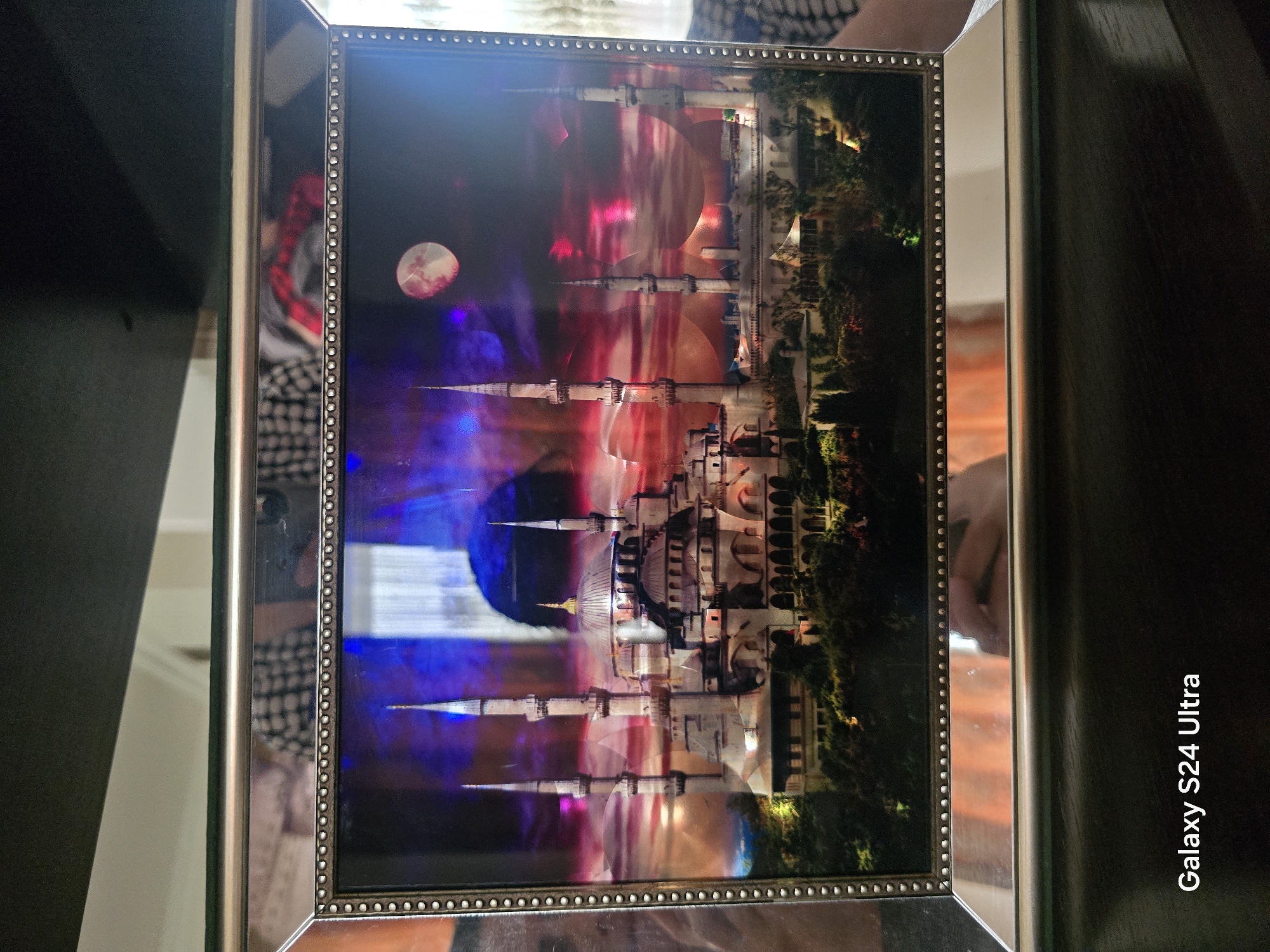 The image shows a framed picture of the Hagia Sophia mosque in Istanbul, Turkey. The picture depicts the mosque at night with a full moon and a reddish-purple sky. The mosque is illuminated, with its minarets and domes clearly visible. The frame of the picture is silver and has a beaded border. The picture is placed on a shelf, and a reflection of a person in a checkered shirt can be seen in the glass. The text "Galaxy S24 Ultra" is visible in the bottom left corner of the image. The image is taken from a slightly elevated angle, looking down at the framed picture. There is no information about Shali, Russia in the image.