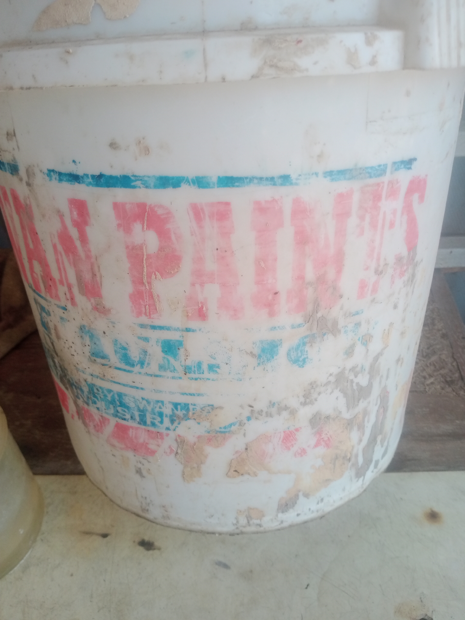 A close-up shot shows the side of a dirty, white plastic bucket with faded pink and blue lettering. The bucket is cylindrical and appears to be empty. The lettering on the bucket is partially obscured by dirt and wear, but "PAINTS" is clearly visible in large pink letters. Below this, a blue stripe runs across the bucket, and beneath that, there is more faded lettering in pink and blue. The bucket is resting on a light-colored surface, possibly a table or counter. The background is blurred and appears to be a wooden surface. This image was taken in Jalingo, Nigeria.