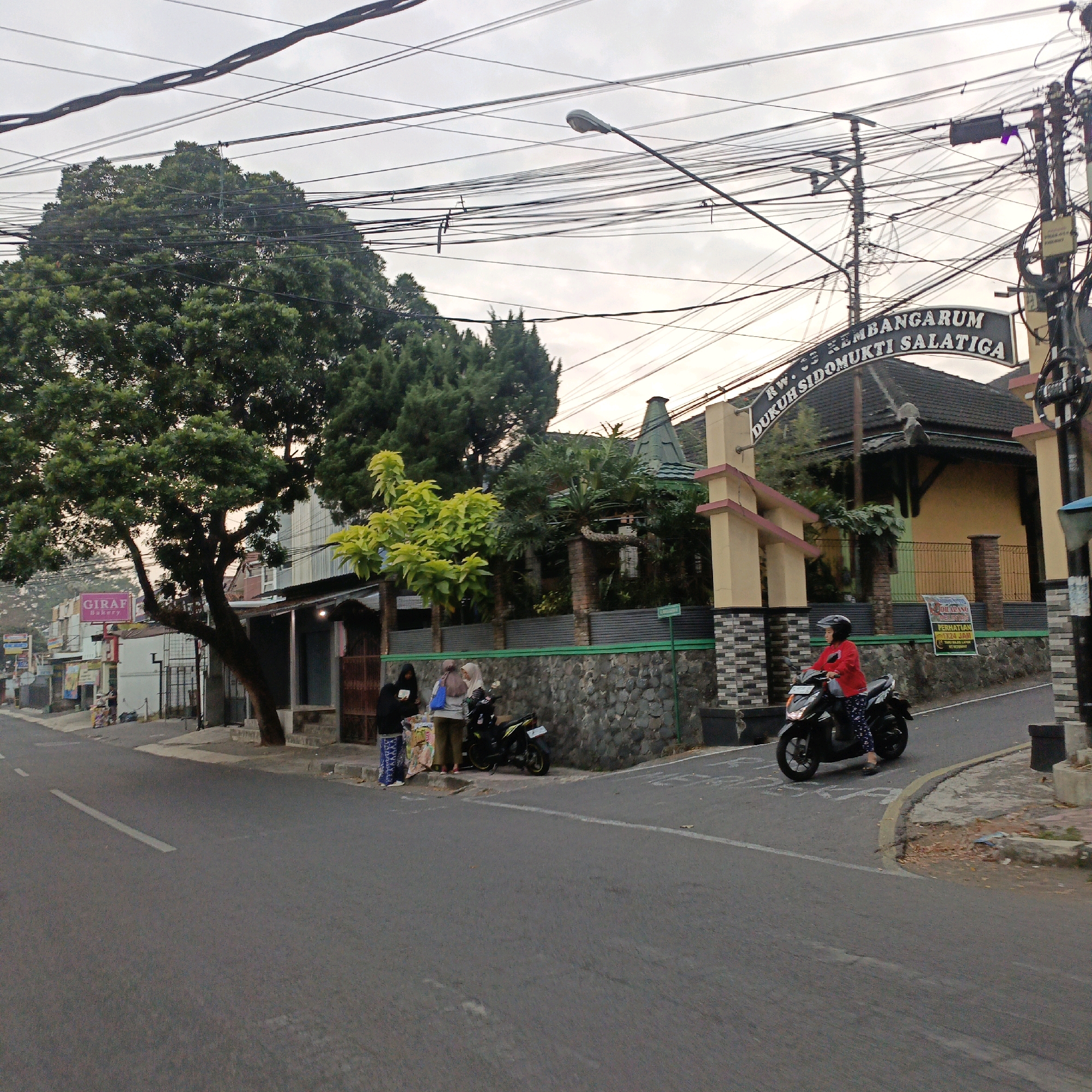 This is a street-level photograph taken in Salatiga, Indonesia. The scene captures a portion of a road in front of a stone-walled structure with a decorative archway above it. The archway is inscribed with text that reads "RW. 03 KEMBANGARUM DUKUH SIDOMUKTI SALATIGA".
In the foreground, a person wearing a red jacket and patterned pants rides a black motorcycle towards the right side of the frame. The motorcycle's headlights are illuminated. On the left side of the street, a few individuals are standing near a small stall or cart. One person is holding a blue bag, and another appears to be looking at items on the cart. A black motorcycle is parked behind them.
The street is paved and has a white lane marking. Beyond the stone wall, there is a building with a dark, angular roof and a yellow exterior. A large tree with dense green foliage is prominent on the left side of the image, casting a shadow. Utility poles and a complex network of overhead electrical wires dominate the upper portion of the frame, suggesting a densely populated urban or suburban area. The sky is overcast with a soft, diffused light. In the background, various buildings and signs are visible, including a sign for "GIRAF BAKERY". The overall impression is of a residential or commercial street in an Indonesian city.