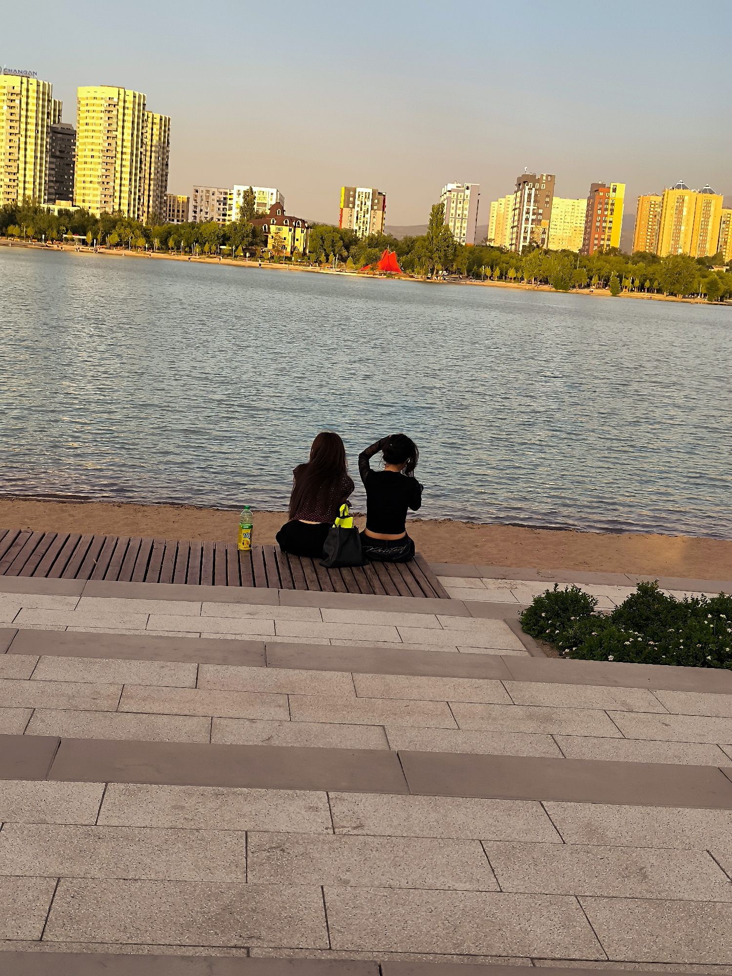 The image depicts a serene waterfront scene in Almaty, Kazakhstan, likely along the Big Almaty Lake or a similar recreational area. In the foreground, two young women are seated on a wooden bench overlooking a calm body of water. One woman, with long dark hair, is wearing a dark patterned top and dark pants. The other woman, also with dark hair, is wearing a black crop top and black pants, with her hair pulled up. Next to them on the bench are a bottle of what appears to be juice and a black bag. The background features a city skyline with several modern high-rise buildings illuminated by the warm glow of the setting sun. Lush green trees line the waterfront, separating the water from the urban landscape. The sky is a clear gradient of pale blue to a soft orange hue, indicative of sunset. The foreground is characterized by a tiered stone pathway, leading down towards the wooden bench and the beach.