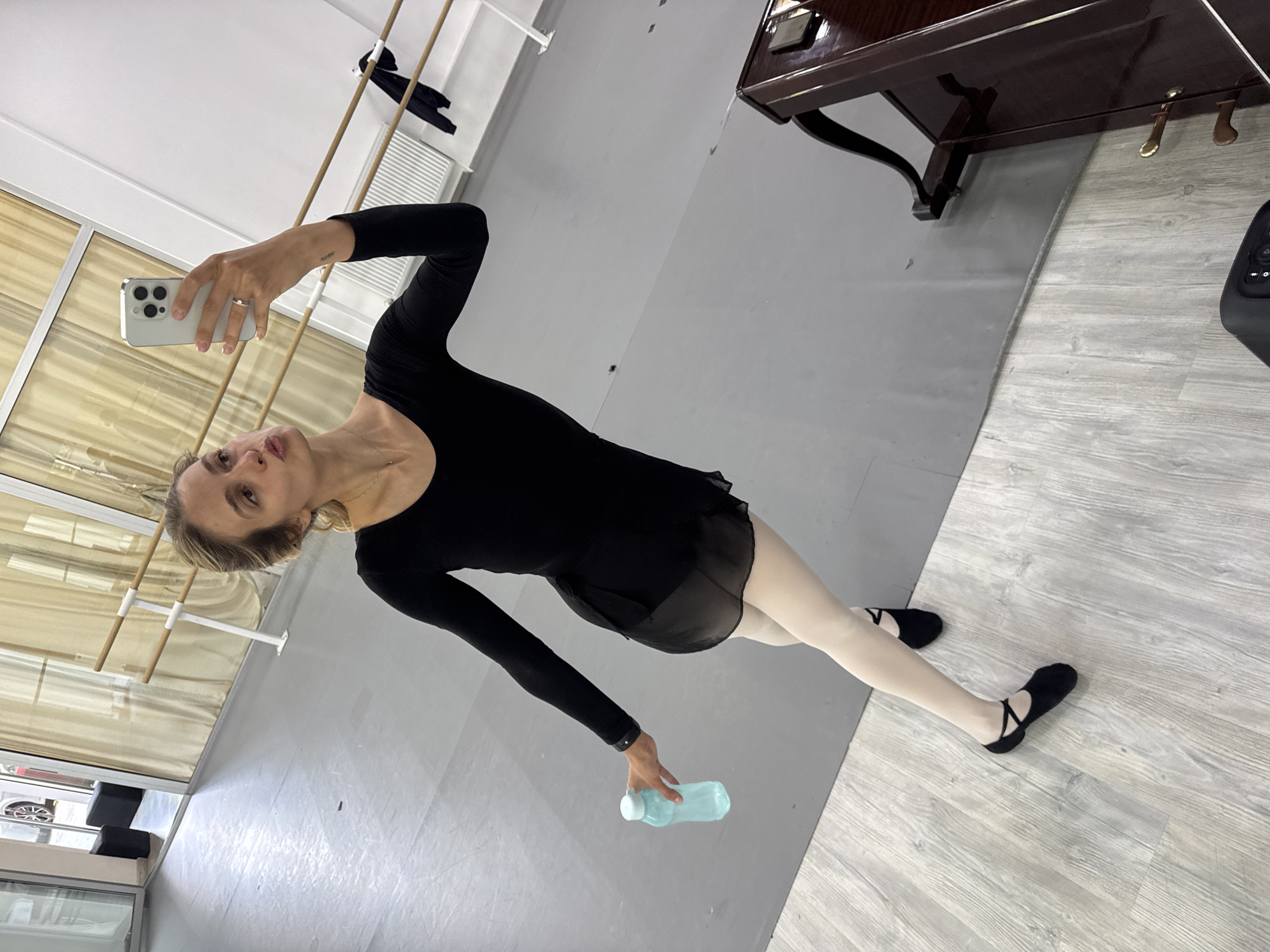 A full-body selfie captures a woman standing in what appears to be a dance studio. She wears a black long-sleeved leotard, a sheer black wrap skirt, light-colored ballet tights, and black ballet shoes. In her right hand, she holds a light-colored smartphone to take the photo, and in her left hand, she holds a light blue water bottle. Her hair is pulled back, revealing a ring on her left hand and a small, dark, indistinct tattoo on her left forearm. Her expression is neutral as she looks towards the camera.
The studio features a light-colored wall with a wooden ballet barre supported by white brackets, visible on the left side, where a black garment hangs. Curtains, appearing light beige or gold, cover windows behind the barre. In the background, to the right, a dark wooden grand piano with brass pedals is partially visible. The floor is divided into two distinct sections: a darker grey, possibly marley flooring, and a lighter grey wood-look floor, creating a diagonal split across the image. The woman stands primarily on the grey flooring, with the wood-look floor extending to the right. Through a window or doorway on the far left, a glimpse of black items and what appears to be a car can be seen outside. The overall lighting suggests an indoor daytime setting.