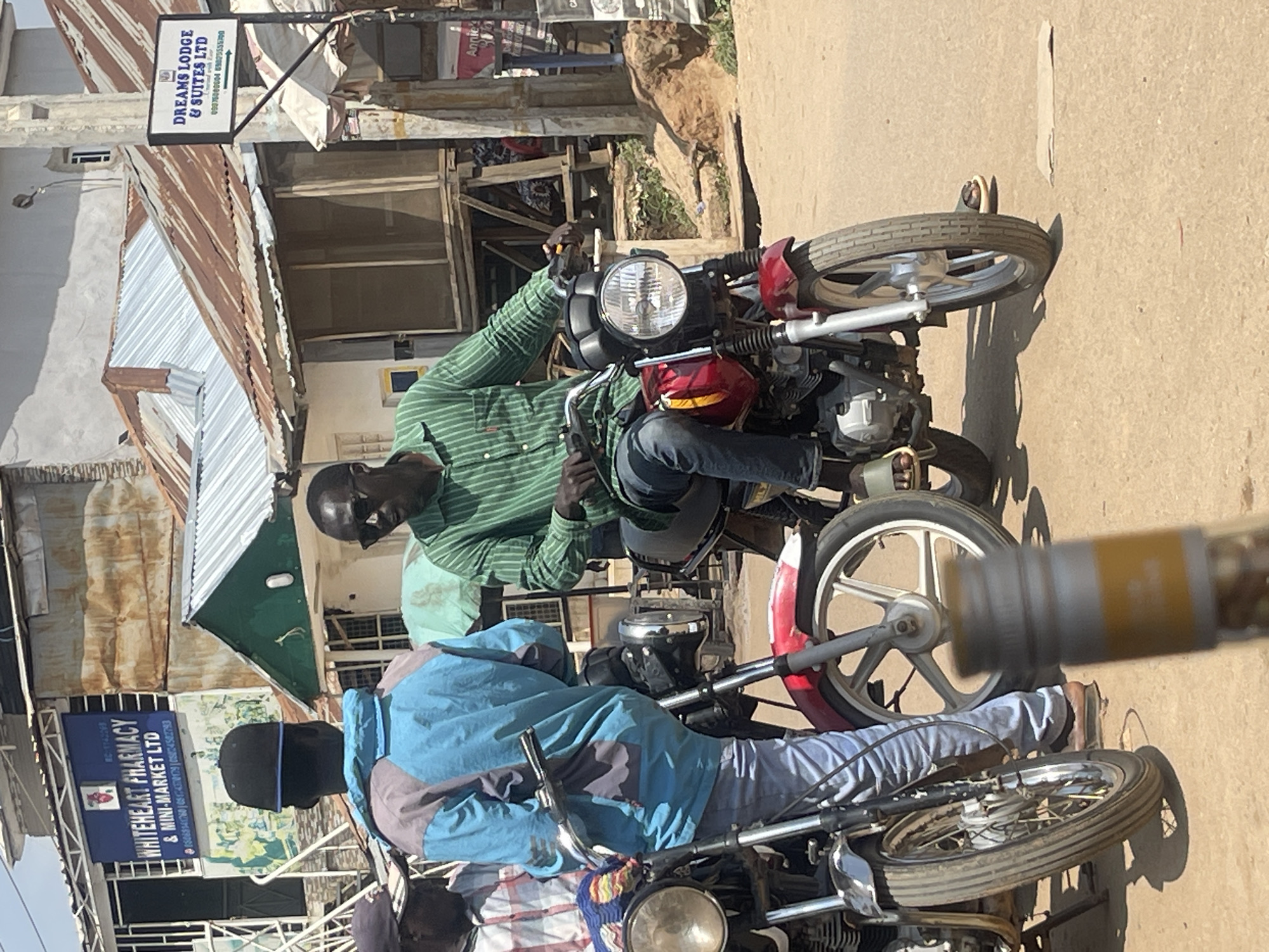A group of men are gathered on a dusty street in Abuja, Nigeria, with motorcycles as their primary mode of transportation. The man in the foreground, wearing a green striped shirt and sunglasses, sits astride a red motorcycle. To his left, another man stands by a motorcycle, wearing a blue and grey jacket and a black cap. Further to the left, a third man in a plaid shirt is partially visible. In the background, buildings with corrugated iron roofs line the street, and a sign for "WHITEHEART PHARMACY & MINI-MARKET LTD" is prominently displayed. Another sign, "DREAMS LODGE & SUITES LTD", is also visible. The environment appears to be a typical urban street scene in Nigeria, with a mix of commercial establishments and residential buildings.