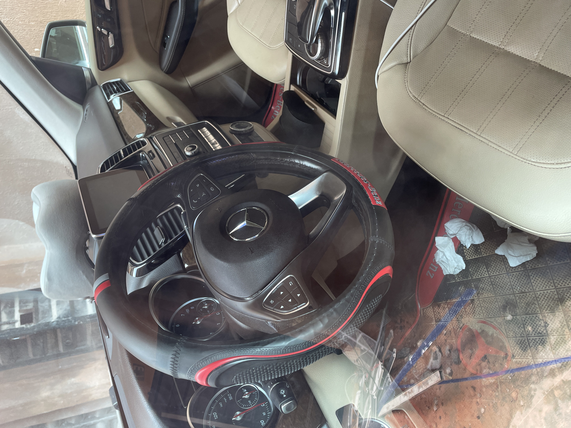 The image shows the interior of a Mercedes-Benz vehicle. The dashboard features a central infotainment screen, air vents, and climate control buttons. The steering wheel is covered in a black and red steering wheel cover and has control buttons. The car has tan leather seats and a tan interior. There are two crumpled white tissues on the floor mat. The floor mat is black and has a red Mercedes-Benz logo. The car appears to be parked on a dirt surface with some debris scattered around. The exterior of the building is visible through the windshield, with brickwork and a light-colored wall. The scene is consistent with the interior of a car parked in a private driveway or lot in Nkpor, Nigeria.