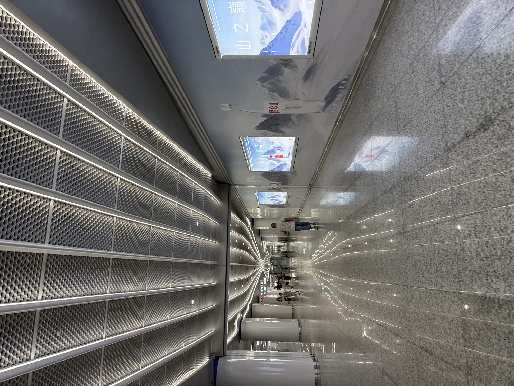 This is a photograph of a well-lit, modern subway station interior in Haizhu District, China. The scene depicts a long, wide concourse with a highly polished, grey tiled floor that reflects the overhead lights and illuminated advertisements. The ceiling is a sophisticated design featuring a honeycomb-like pattern with integrated LED strip lighting that follows a curved architectural line, creating a sense of depth and perspective.
On the right wall, a large, continuous advertisement features a snowy mountain landscape with a prominent image of a water bottle labeled "昆仑山" (Kunlun Mountain). Several smaller illuminated panels within this advertisement display similar mountain imagery and Chinese text.
Several individuals are present in the concourse, walking in various directions. In the foreground, a man wearing a striped shirt and jeans walks away from the camera. Further down the concourse, a group of people, including adults and at least one child, are walking towards the camera. The overall atmosphere is clean, spacious, and contemporary, indicative of a functional and aesthetically designed public transportation hub. The lighting is bright and consistent, illuminating the space effectively.