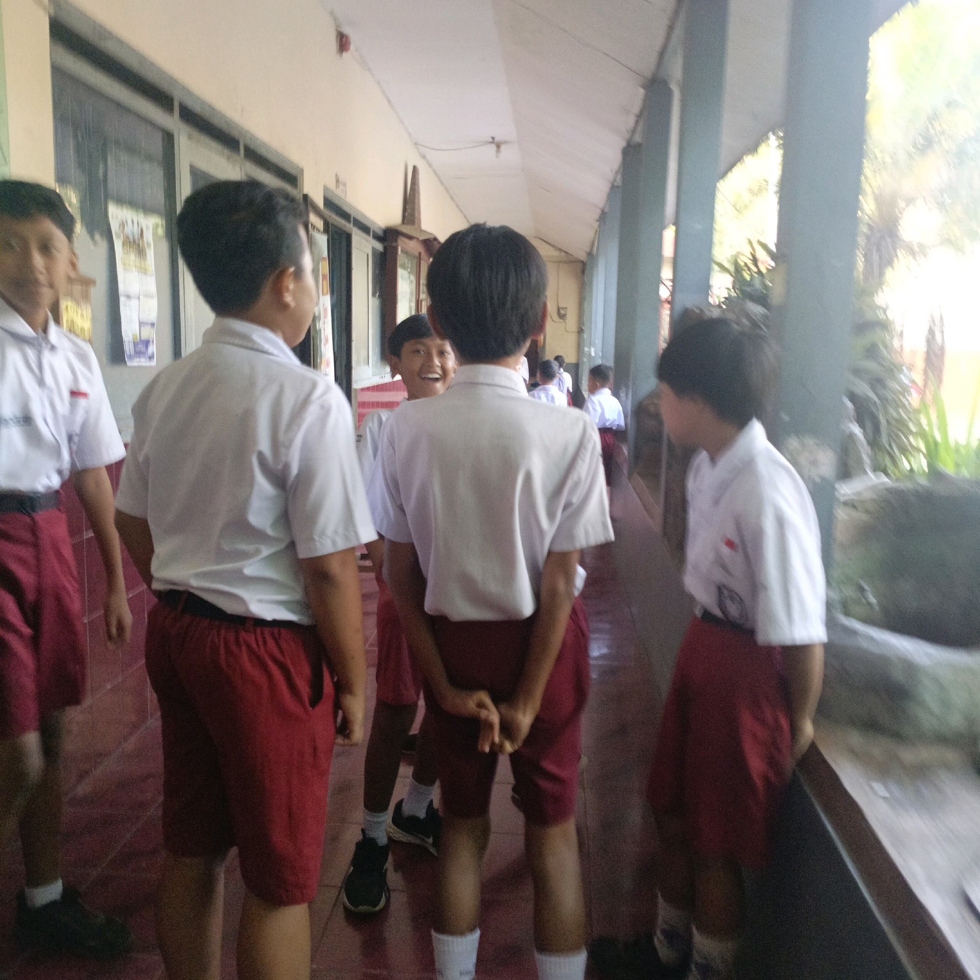 In Salatiga, Indonesia, the image captures a group of male schoolchildren in white shirts and red shorts standing in a covered walkway. The walkway appears to be part of a school building, with windows and a tiled floor visible. Sunlight streams in from the right, illuminating the scene and casting shadows. The children are in various poses, some looking at the camera, others conversing. Their white shirts have short sleeves and collars, and some have patches or logos on the chest. Their red shorts are knee-length. The walkway has teal-colored columns and a beige roof. In the background, there are hints of vegetation and what appears to be a stone feature. The overall impression is of a casual, everyday moment at a school.