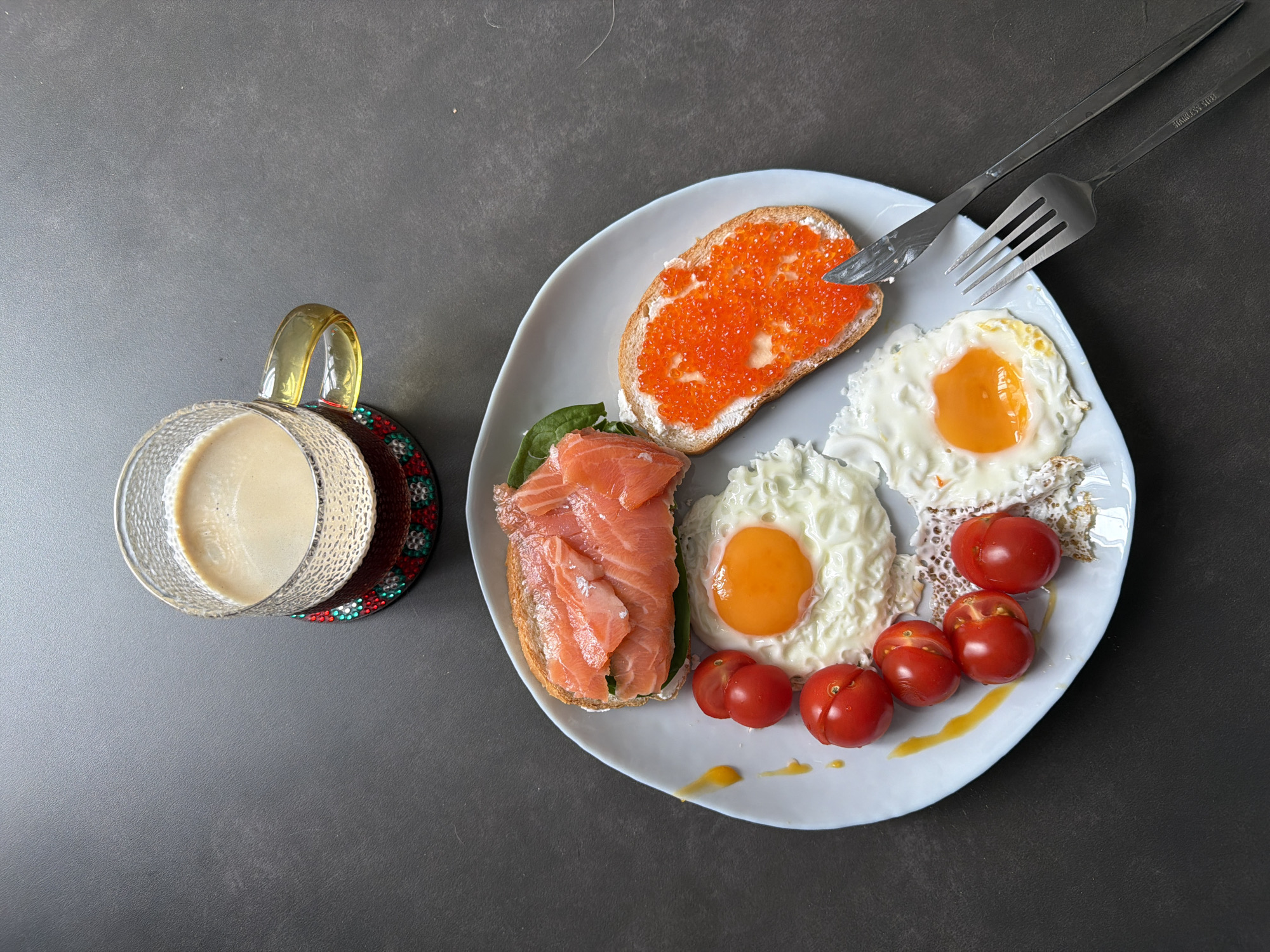 The image shows a breakfast scene on a dark gray surface. A clear glass mug filled with a light-colored beverage sits to the left of a white plate. The plate contains a slice of toast topped with red caviar, a sandwich with smoked salmon and greens, two fried eggs, and several small red tomatoes. A knife and fork rest on the upper right side of the plate. The scene does not suggest a specific location in the city of Unknown, Unknown.