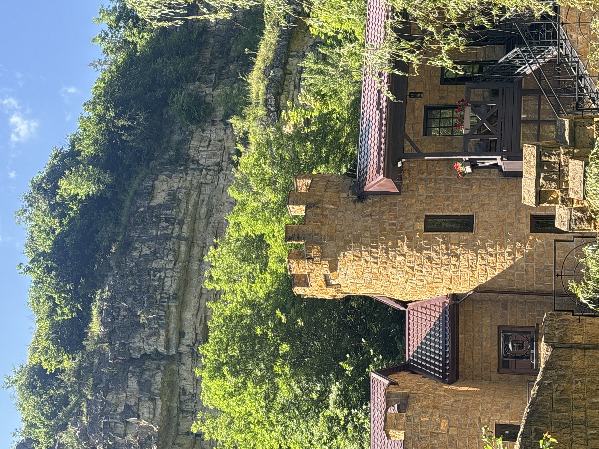 This media file captures a sunny day scene in Kislovodsk, Russia, featuring a stone building with a castle-like turret and a dramatic, tree-lined cliff face. The building's exterior is constructed from light brown, textured stone tiles, and it features brown tiled roofs and dark metal railings. A black metal staircase ascends along the side of the building. Red flowers in hanging planters add touches of color. Lush green trees surround the building and cling to the rocky cliff, which displays layers of sedimentary rock formations. The sky is bright blue with a few scattered white clouds. The sun casts shadows, indicating it is likely midday. The overall impression is one of a serene and picturesque natural and architectural setting. There is no visible text in the image.