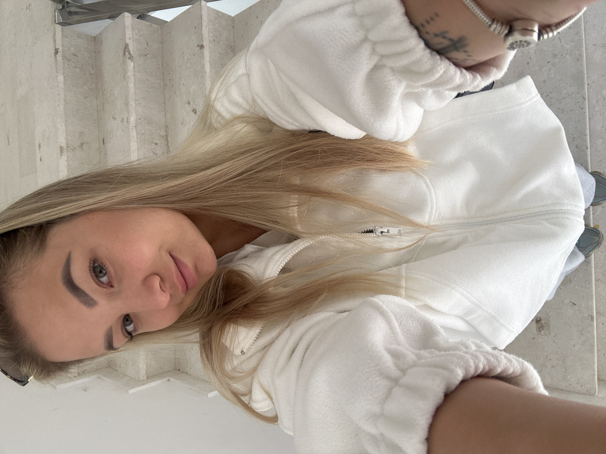A woman with blonde hair and green eyes is standing on a set of stone stairs, possibly in the city of Budva, Montenegro, based on the stone architecture common to the area. She is wearing a white fleece jacket and has a silver bracelet on her wrist. The woman is holding the camera above her and looking down at it, creating a selfie perspective. The background includes stone stairs with a polished metal handrail on the left side. Her shoe appears to be gray. The woman is wearing sunglasses in her hair.