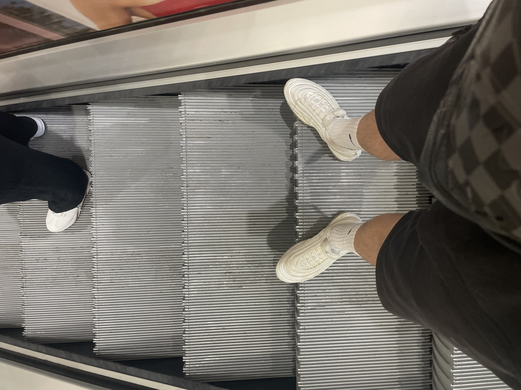 The image depicts the feet of two individuals on an escalator. On the left, a person is wearing black pants and white sneakers. On the right, another person is wearing dark shorts, white socks, and white sneakers. The escalator steps are grey. The photograph does not provide enough information to determine the specific location in Sofia, Bulgaria.