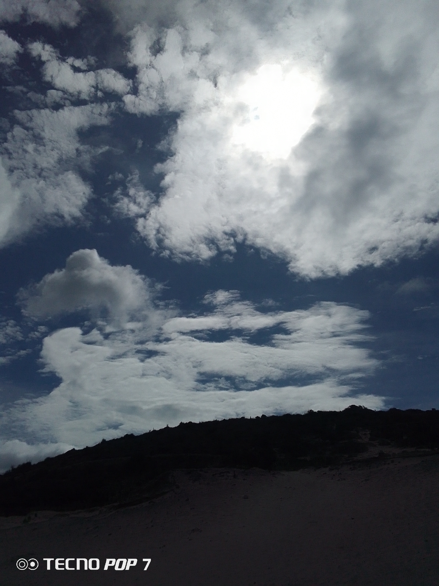 The photograph depicts a landscape in Pedro González, Venezuela. The bottom third of the image is dominated by a dark, sandy terrain, possibly a beach or dune. Above this, a silhouette of a low-lying hill or ridge is visible. The sky occupies the majority of the frame, with a mix of dark blue and white clouds. A bright, diffused light source, likely the sun, is partially obscured by the clouds. The text "TECNO POP 7" is present in the bottom left corner of the image.