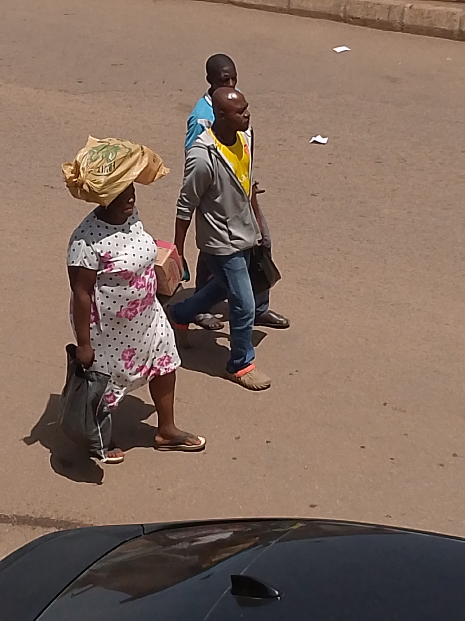 A medium shot captures three individuals walking across a dusty, light brown asphalt surface. In the foreground, a woman is walking from left to right, carrying a large, light brown plastic bag filled with produce on her head. She wears a white dress with black polka dots and a pink floral pattern. She is also holding a black plastic bag in her left hand. To her right, a man in a grey hooded jacket and blue jeans walks past, carrying a black bag in his right hand. Behind him, another man in a blue shirt walks. The scene appears to be on a street in a city, with a curb made of stone blocks visible in the upper portion of the image. Two white pieces of paper are scattered on the ground. A dark colored car is partially visible in the bottom of the frame.