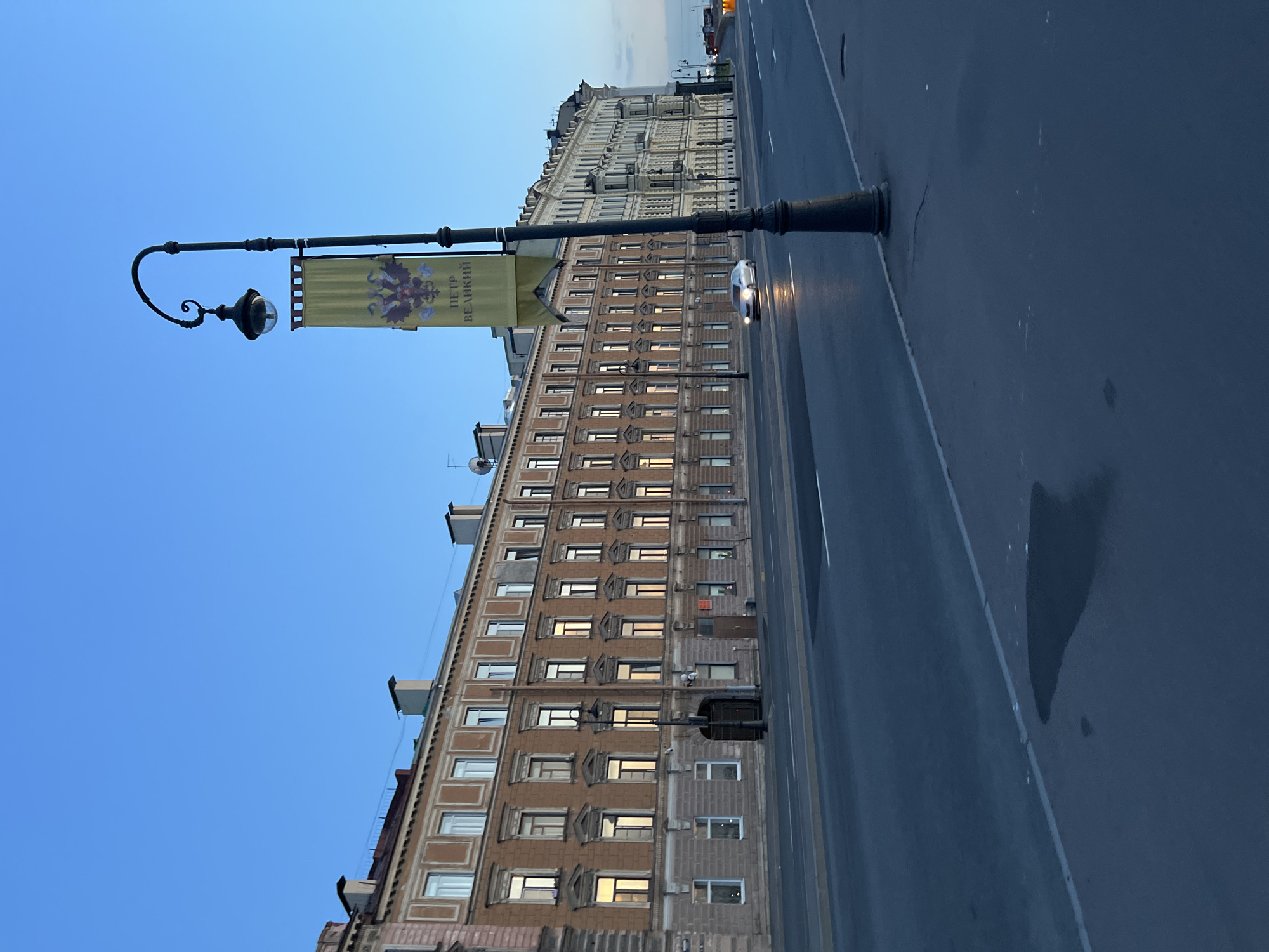 The image captures an urban scene at dusk in Saint Petersburg, Russia. A long, multi-story building with numerous windows, some of which are illuminated, lines a wide, empty street. A vintage-style lamppost with a decorative banner featuring an eagle and Russian text stands on the sidewalk in the foreground. A silver car is seen driving on the street, its headlights reflecting on the wet asphalt. The sky is a deep blue, indicating twilight. The architecture suggests a historic European city.