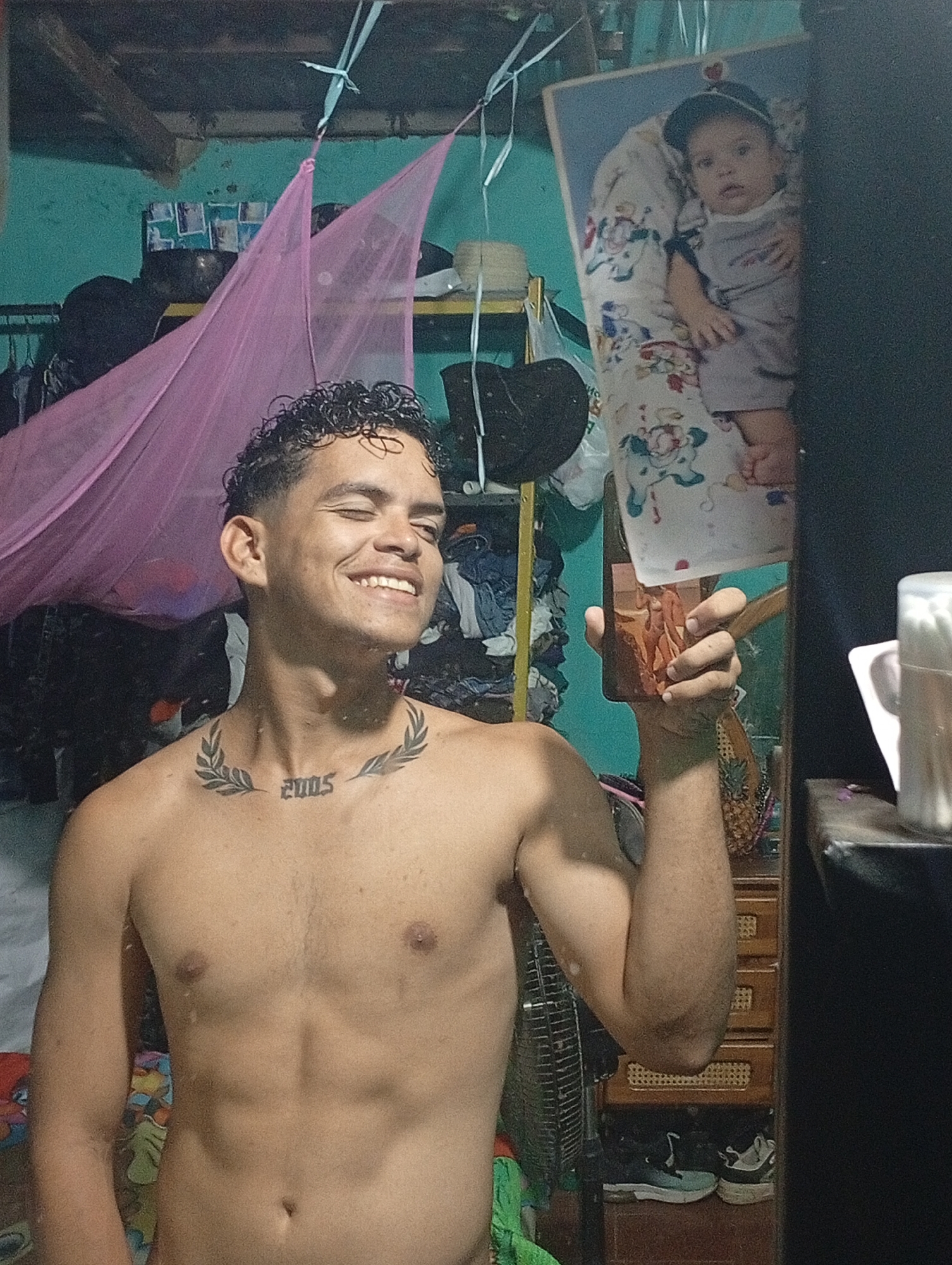 A shirtless young man with dark, curly hair and a tattoo of "2005" surrounded by laurel leaves on his chest is smiling at the camera, which appears to be a phone he is holding in his right hand. The photo is likely taken in a bedroom in Altagracia, Venezuela. The background includes a pink mosquito net, a cluttered shelf with clothes and other items, and a framed photograph of a baby. The reflection in the mirror reveals a wooden dresser, sneakers on the floor, a fan, and a container of cotton swabs. The location is assumed to be in Altagracia, Venezuela based on the prompt.