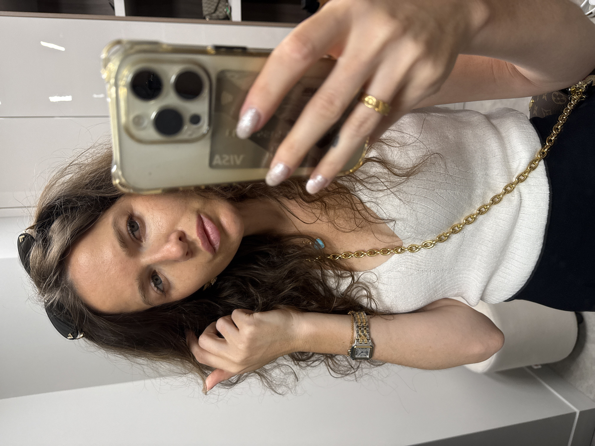 A woman takes a selfie in a room, possibly a dressing room or closet, given the presence of clothing racks and drawers in the background. She is wearing a white ribbed tank top, black pants, and a gold chain necklace with a turquoise pendant. On her left wrist, she wears a two-tone watch, and on her right ring finger, a gold ring. Her nails are painted with a pearlescent polish. She is holding a gold-encased iPhone in her right hand, which is reflecting light from above. Sunglasses are perched on top of her head. The background is primarily white and grey with some visible shelving and what appears to be a white stool. The image is well-lit and the woman's facial features are clearly visible, though the focus is slightly soft.