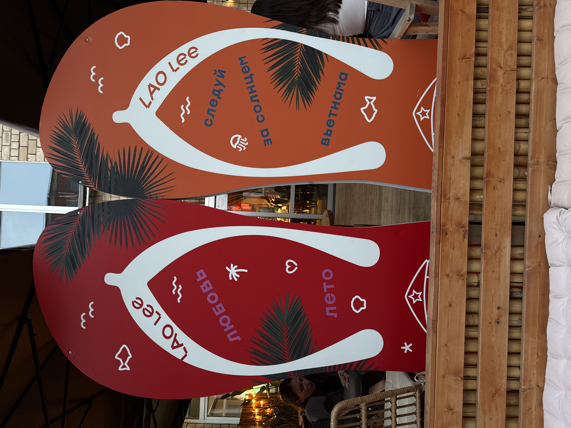 This image shows two large, vertically aligned, sandal-shaped advertisements for a brand called "Lao Lee." The top sandal is orange, and the bottom sandal is red. Each sandal features white straps and Russian text, as well as graphics like palm leaves, clouds, and stylized icons. The text on the orange sandal includes the phrases "следуй за солнцем" (follow the sun) and "Вьетнама" (Vietnam). The text on the red sandal includes the phrases "любовь" (love) and "лето" (summer).
The sandals are mounted on a wooden structure, possibly a divider or decorative element in an outdoor or semi-outdoor space. In the background, we see hints of a brick building, a window, and other furniture. People are partially visible in the image, mostly blurred in the background, suggesting this is a public area.
The sandals are a promotion for "Lao Lee" located in Moscow, Russia.