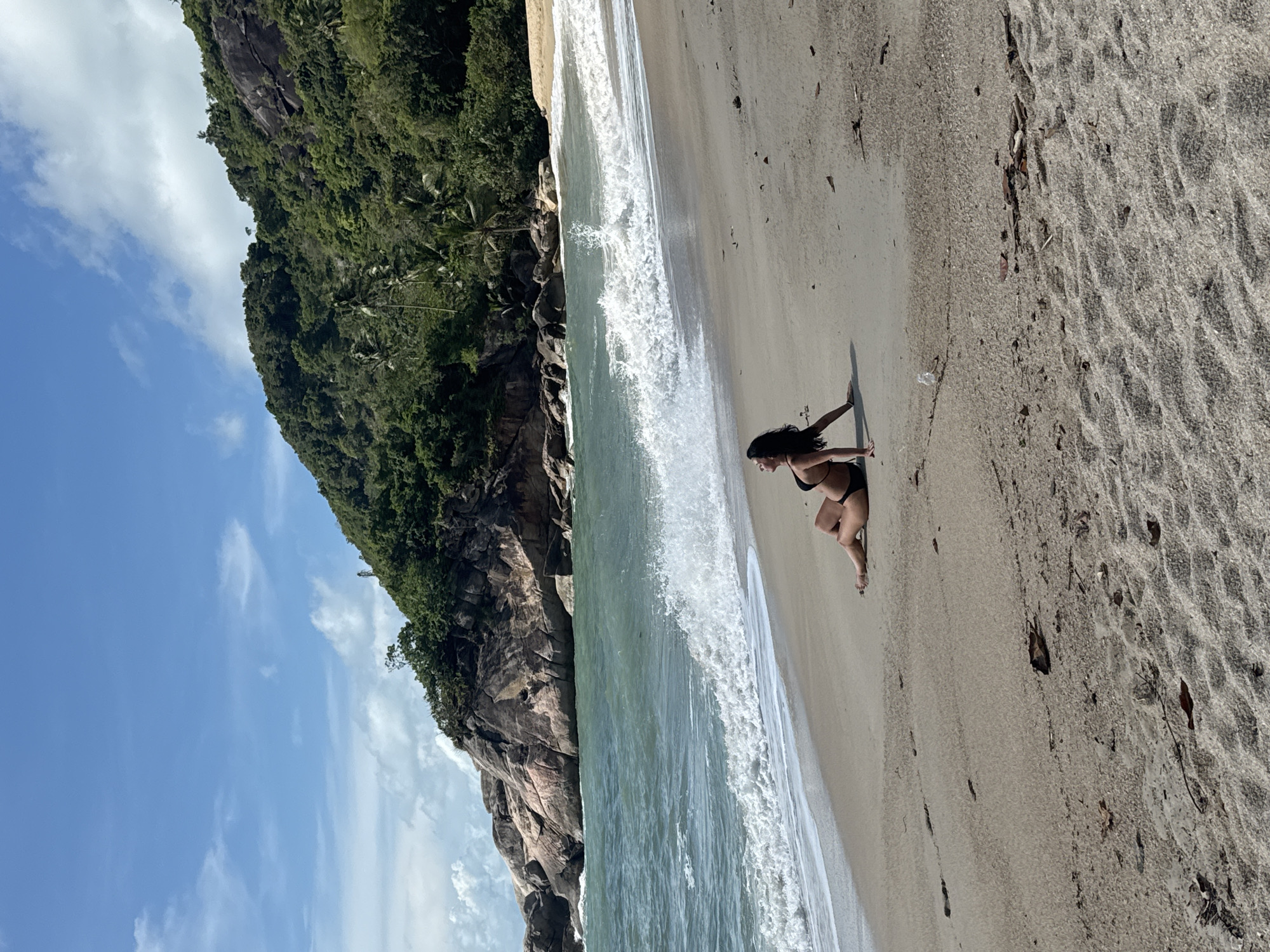 A medium shot depicts a woman sitting on a sandy beach in Baan Thong Nang, Thailand. She is wearing a black bikini. Her hair is long and dark. She is facing the ocean, her arms extended to her sides. The surf is visible and gently rolling onto the shore. The beach is sandy and contains debris such as leaves and twigs. To the left, there is a rocky cliff covered in dense green vegetation. The sky is blue with scattered clouds.