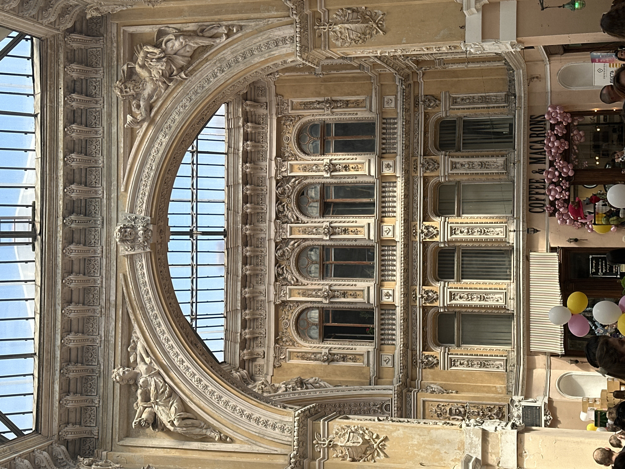 The image displays an interior view of a grand, multi-story architectural passage in Odesa, Odesa Oblast, Ukraine, likely within "The Passage" building. The scene is illuminated by natural light filtering through a large, arched glass and metal-framed skylight at the top. Below the skylight, an ornate, weathered light yellow facade is visible, featuring a prominent central arch. Resting on the spandrels of this arch are two classical-style reclining sculptures: a female figure holding an object on the left, and a bearded male figure with a quiver on his back and holding an object on the right. Above the arch, an elaborate frieze with decorative brackets and carvings runs horizontally.
Within the central arch, a section of the building's upper floor is visible, featuring multiple arched windows that reflect the sky. Below this, the facade continues with two levels of intricately decorated windows. These windows are framed by pilasters and various classical ornaments including masks, garlands, and small caryatids. Some windows on the upper level are open, while those on the lower level are either closed or covered by internal blinds.
At ground level, the passage hosts commercial establishments. On the right, a shop named "COFFEE & MACARONS" has its entrance adorned with a large cluster of pink balloons forming an archway. Inside, a red hat is visible on display. Adjacent to this shop, a vertical sign partially displays "УКРОРОНА ЯКІСТЬ" (Ukroborona Yakist), indicating "Ukrainian Defense Quality." On the left side of the passage, a segment of another shop or entrance is visible, featuring a striped awning (light pink and white) and a gathering of white and yellow balloons. Numerous individuals, mostly blurred, are visible from the head and torso down in the foreground at the bottom of the image, indicating a crowd present in the passage. The architectural elements throughout the passage, including columns and walls, are embellished with carved coats of arms or emblems. The overall color palette is dominated by the light yellow/beige of the facade, with accents of white, pink, and black.