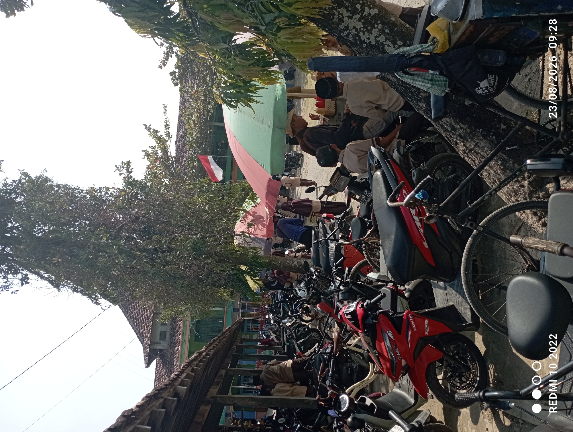 The image depicts a busy outdoor scene in Dukuhseti, Indonesia, likely a schoolyard or marketplace, given the presence of many parked motorcycles and bicycles. A building with a tiled roof and green accents is visible in the background. Several people are present, including children in traditional Islamic attire and adults. Some individuals are gathered around food stalls shaded by large umbrellas. A man wearing a hat appears to be serving food, interacting with a group of children. A red and white Indonesian flag is flying from a pole. The environment is sunny, with trees providing shade. The foreground features a variety of parked motorcycles and bicycles, indicating a gathering of people. Some of the motorcycles have visible license plates. The overall atmosphere suggests a lively community event or daily activity.