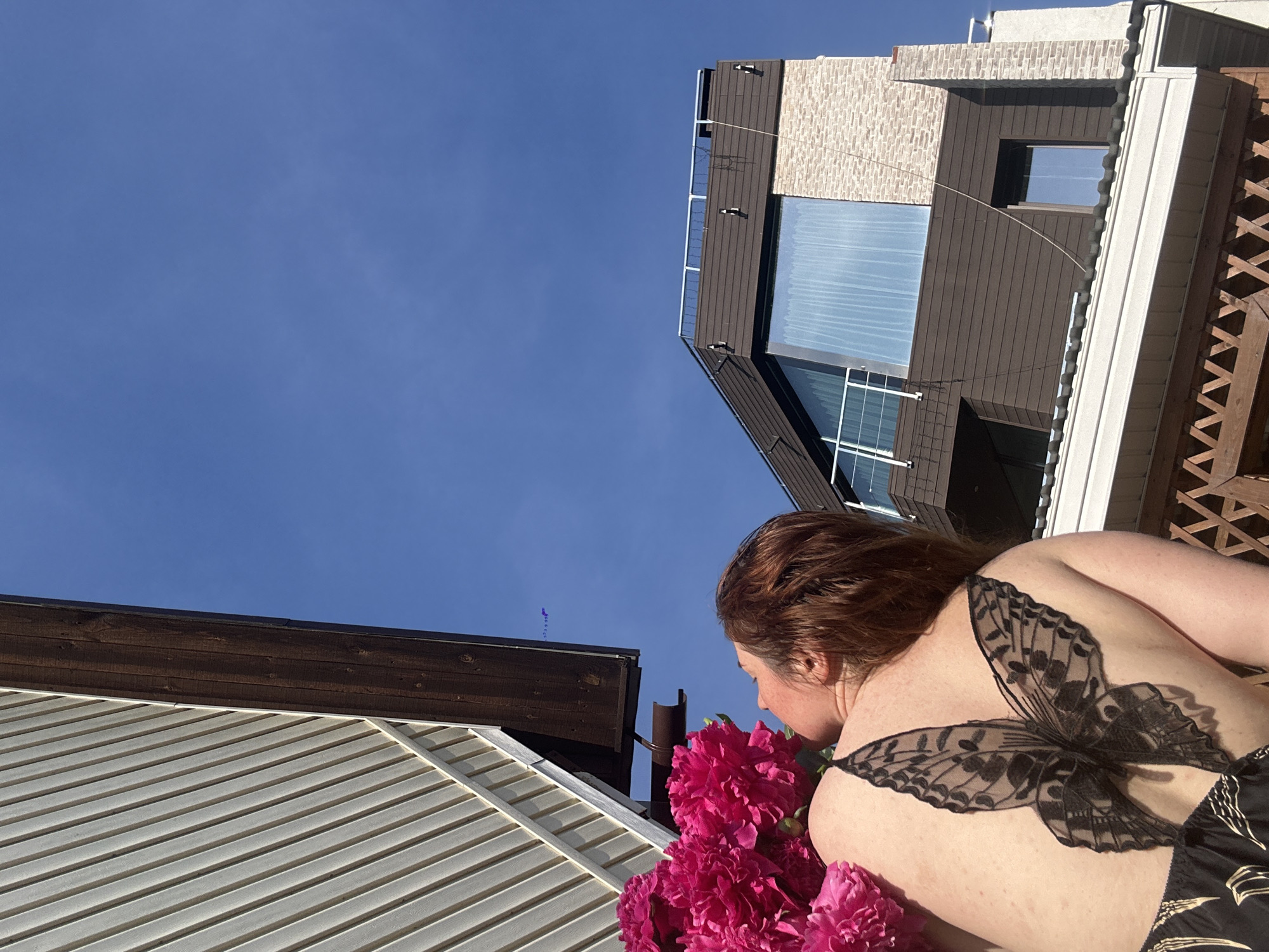 A woman with auburn hair and freckles is shown from the back, smelling a bouquet of bright pink flowers. She is wearing a black garment with a lace butterfly design on her upper back. The scene takes place outdoors, with a clear blue sky above. In the background, modern architectural structures with dark brown siding and brick accents are visible, suggesting an urban or suburban setting. The buildings appear to be multi-story with windows and balconies. One building has a large, angled window on its upper level. The overall impression is of a sunny day. The location is Nizhny Novgorod, Russia.