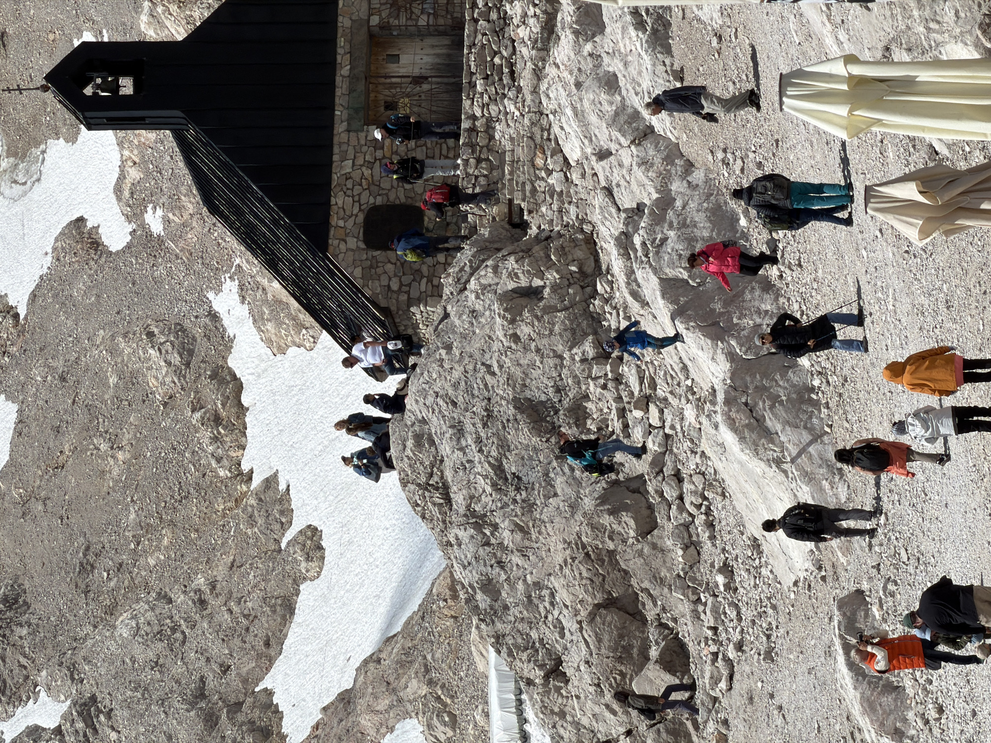 The image depicts a mountainous landscape near Garmisch-Partenkirchen, Germany, with a small stone building topped by a dark, steep-roofed structure resembling a chapel or a mountain hut. The terrain is rocky and rugged, with patches of snow visible on the slopes. Several people are scattered throughout the scene, engaged in various activities.
In the foreground, on the left, a person in an orange vest is taking a photograph. Beside them, a person in a black jacket and hat is standing. Further to the right, a group of people are walking and standing on the rocky ground. One person in a pink jacket is climbing. Another person, wearing a black jacket and teal pants, is standing near a backpack.
In the middle ground, a group of people are ascending a stone staircase leading up to the building. Some are carrying backpacks, and one individual is wearing a red backpack and using trekking poles.
In the background, on a higher rocky outcrop, a cluster of people are gathered, looking towards the building. The building itself has a stone facade with a wooden door and a cross atop the dark roof. The surrounding environment is characterized by steep, rocky inclines and sparse vegetation. The sky is bright, suggesting a sunny day. Several folded umbrellas are visible in the lower right corner, suggesting a possible outdoor seating area.