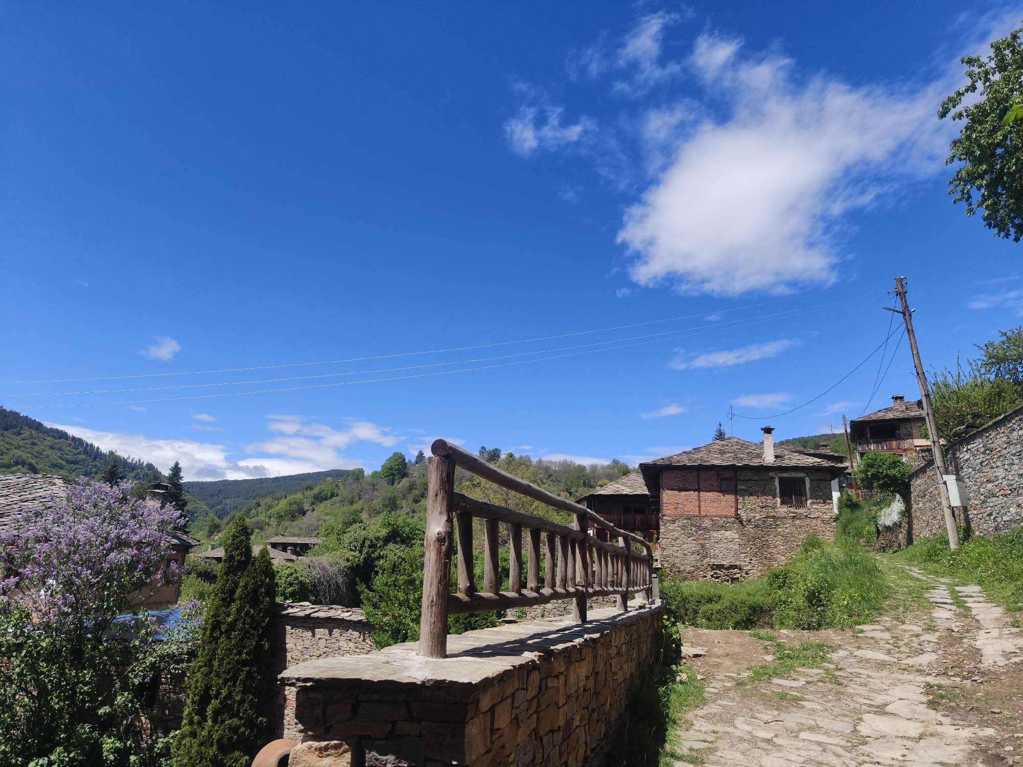 The image captures a sunlit scene in Kovachevitsa, Bulgaria. In the foreground, a rustic wooden fence tops a stone wall. Behind the fence, a stone and brick building with a slate roof stands. The path to the right is made of rough, uneven stone, flanked by a stone wall and a telephone pole. In the distance, wooded hills rise under a bright blue sky dotted with scattered clouds. A flowering tree with purple blooms adds a splash of color to the left side of the image.