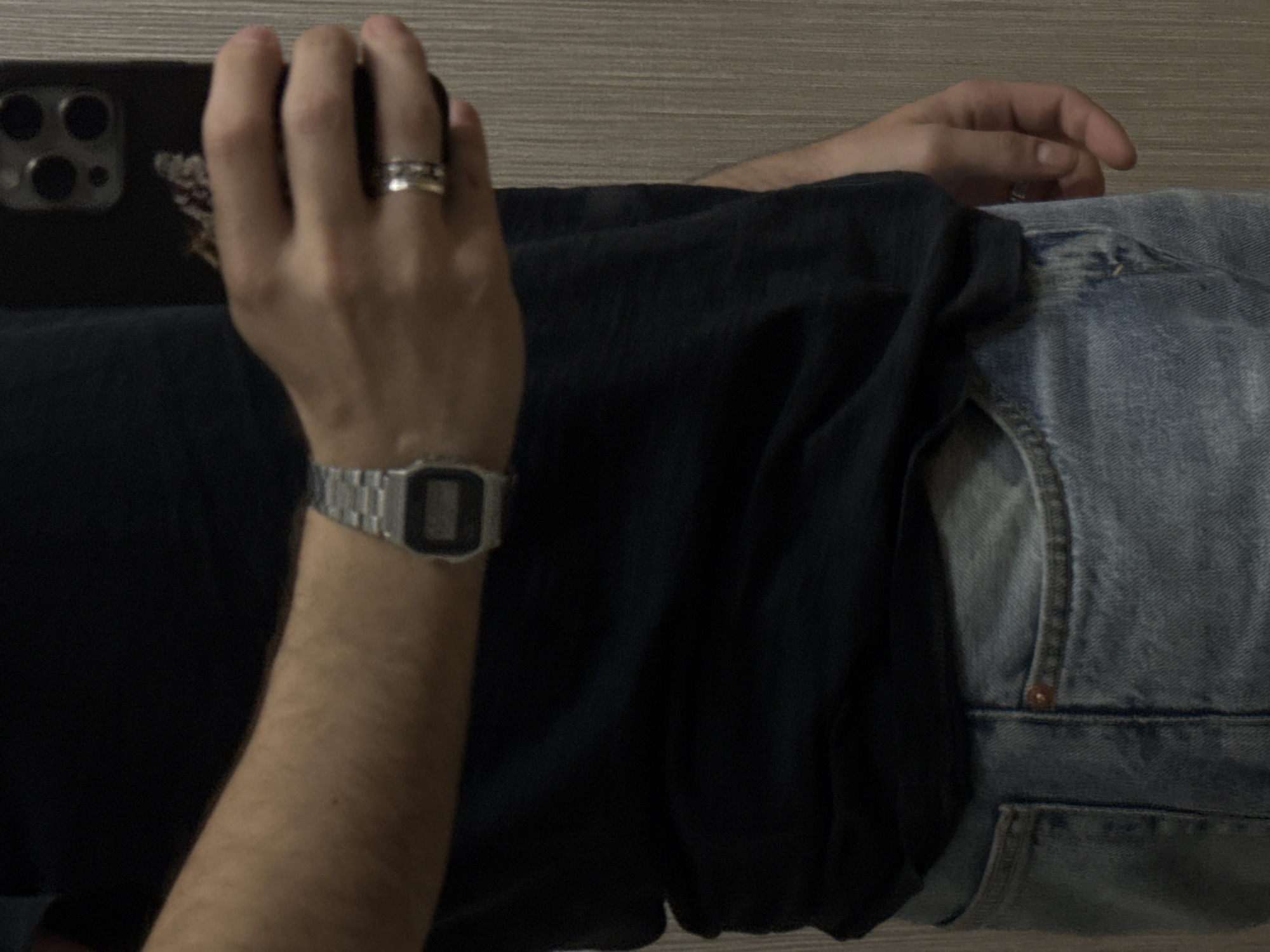 The image displays a partial view of an individual, from the chest to the upper thighs, against a uniformly textured light brown or beige wall with vertical striations. The person is wearing a dark, possibly black, short-sleeved t-shirt and light blue denim jeans. The left arm is raised, with the hand holding a dark-colored smartphone, likely reflecting the scene as if taking a mirror selfie. A silver-toned digital wrist watch with a black display is visible on the left wrist. A silver-toned ring, appearing as two stacked bands, is worn on the left ring finger. The smartphone's rear casing is visible, featuring a triple-lens camera system and a flash. A faint pattern is discernible on the lower back of the phone case. The specific location within the city of Yerevan, Armenia, cannot be determined from the visual information provided.