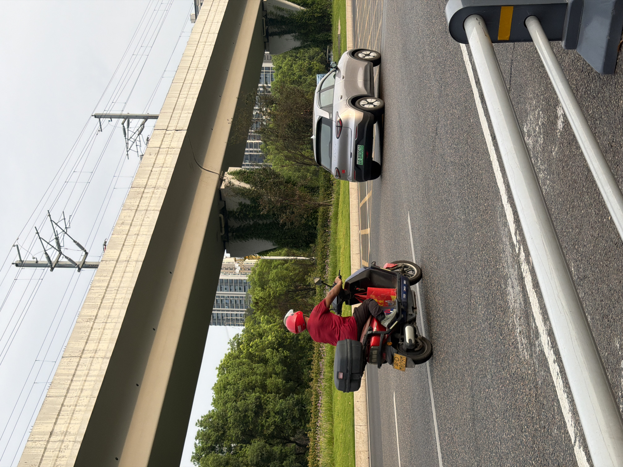 In Jiangjin, China, on a clear day, a person wearing a red shirt and a red helmet rides a red electric scooter down a paved road. The scooter has a black cargo box on the back and a red shopping bag hanging on the side. A silver car is driving in the next lane. Above the road, a concrete overpass supports train tracks and electrical wires. Green trees and tall buildings are visible in the background. A white guardrail with yellow accents is in the foreground.